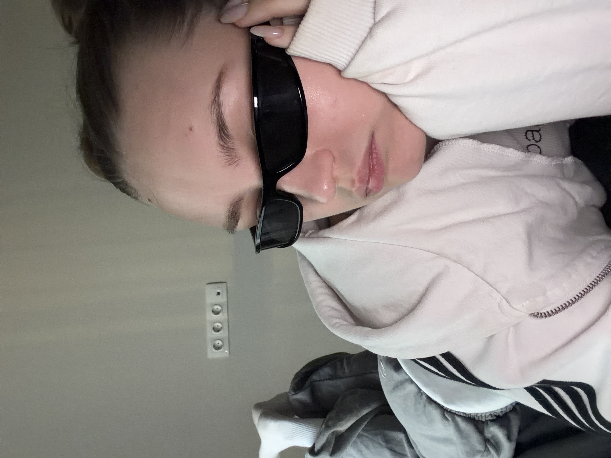 A Caucasian woman with light skin and dark hair pulled back in a bun is resting her head on her hand. She is wearing a light-colored hooded sweatshirt and black sunglasses. In the background is a gray wall with a white electrical outlet. The image does not contain enough information to determine the location in Moscow, Russia.