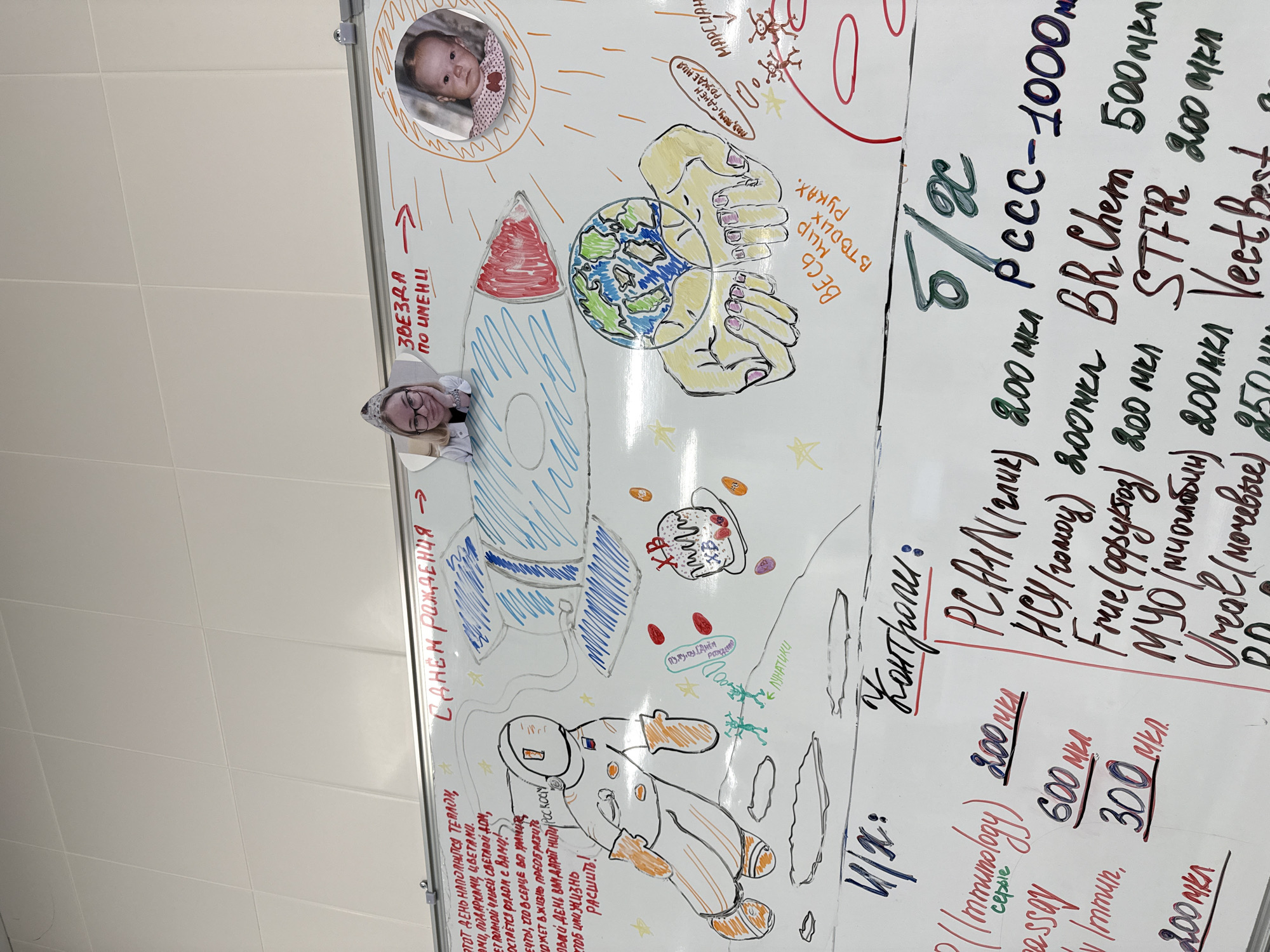 This image displays a decorated whiteboard in an indoor setting, likely a professional or institutional environment in Moscow, Russia. A woman with blonde hair and glasses is visible peeking over the top edge of the whiteboard, looking at the camera with a neutral expression.
The whiteboard is covered with colorful drawings and text, celebrating a birthday with a space theme. Central to the display are a large blue and white rocket, an astronaut in an orange and white suit, and a depiction of the Earth held within two hands, accompanied by the Russian text "ВЕСЬ МИР В ТВОИХ РУКАХ" (The whole world in your hands). A circular photo of a baby is attached near the top right, styled as the sun, with the words "ЗВЕЗДА ПО ИМЕНИ" (Star by the name of...). Other whimsical space elements include drawings labeled "МАРСИАНЕ" (Martians) and "ЛУНАТИКИ" (Lunatics).
Birthday greetings of "С ДНЕМ РОЖДЕНИЯ" (Happy Birthday) are prominently featured, along with a drawing of a birthday cake. A heartfelt poem written in Russian on the lower left side begins with "ЭТОТ ДЕНЬ НАПОЛНИТСЯ ТЕПЛОМ..." (This day will be filled with warmth...) and conveys wishes for happiness, gifts, and the fulfillment of dreams.
A notable section on the right side of the whiteboard contains what appears to be a list of medical or laboratory test parameters under the headings "И/К:" and "Контроли:" (Controls:). This list includes abbreviations like "PCAAN", "HCY", "BR Chem", "STFR", "Frue (фруктоз)", "МУО (миоглобин)", "Urea (мочевые)", and "Veet Best", each followed by numerical values, often expressed in "мкл" (microliters). This detail suggests the celebration may be taking place in a medical or scientific facility. The white ceiling tiles visible above the board further confirm an indoor, possibly institutional, setting.