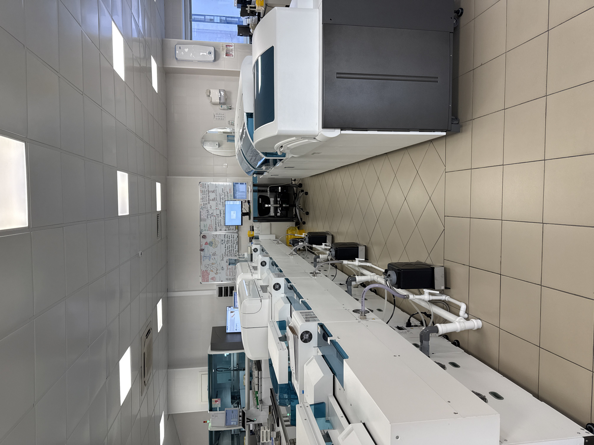 The image displays a clean, well-lit laboratory environment in Moscow, Russia, equipped with an extensive array of automated diagnostic machinery. No individuals are present in the scene.
The primary subject is a long, modular system of interconnected laboratory equipment, colored predominantly white with some dark grey and light blue components. This sophisticated apparatus, likely an automated clinical chemistry or immunoassay analyzer, extends across the room, suggesting a high-throughput processing line for samples. Various pipes, hoses, and electrical conduits are neatly routed along the back wall, connecting to the machinery.
Behind the equipment, on the left, a workstation is visible with two computer monitors displaying software interfaces or data, accompanied by an office chair. A large whiteboard directly behind this station is covered with handwritten notes, diagrams, and text, including what appears to be Russian Cyrillic script with terms like "схема" (scheme/diagram), "показатели" (indicators), and "данные" (data), along with numerical entries like "6/10".
The room features light-colored tiled walls and a light beige tiled floor, laid in both square and diagonal patterns. The ceiling is composed of a grid of white panels interspersed with bright, rectangular fluorescent light fixtures, providing ample illumination. Through a window in the background, another building exterior is partially visible, indicating that it is daytime. Additional wall-mounted fixtures include small dispensers or sinks and a circular mirror. The overall impression is that of a modern, professional clinical or research laboratory designed for automated analytical processes.