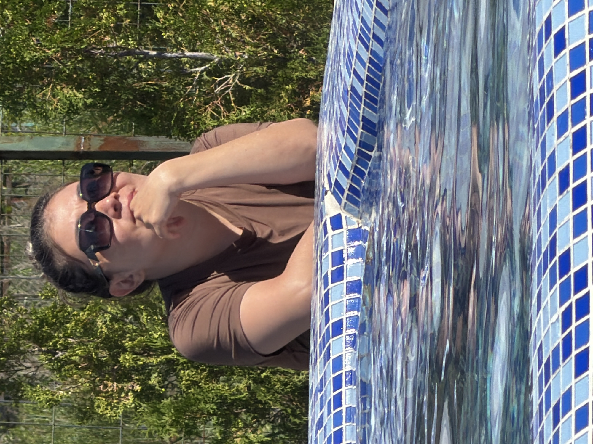 A woman wearing sunglasses and a brown t-shirt leans on the edge of a swimming pool, her chin resting on her hand. She is looking to her right. The pool is lined with blue and white square tiles, and the water is rippling. Behind her, a green bush and a fence are visible. The image is taken outdoors on a sunny day.