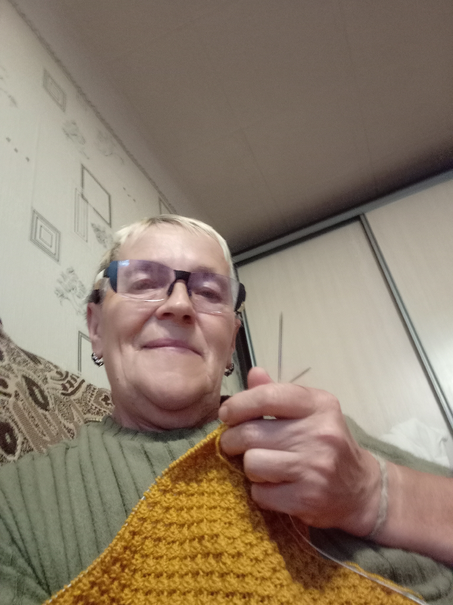 A woman is knitting a yellow garment in a room. She has short blonde hair, wears glasses and hoop earrings, and is dressed in a green sweater. The room's walls are covered in wallpaper with a geometric pattern. To the right of the woman, a closet door with sliding panels is visible. The woman appears to be in her home in the city of Degtyarsk, Russia.