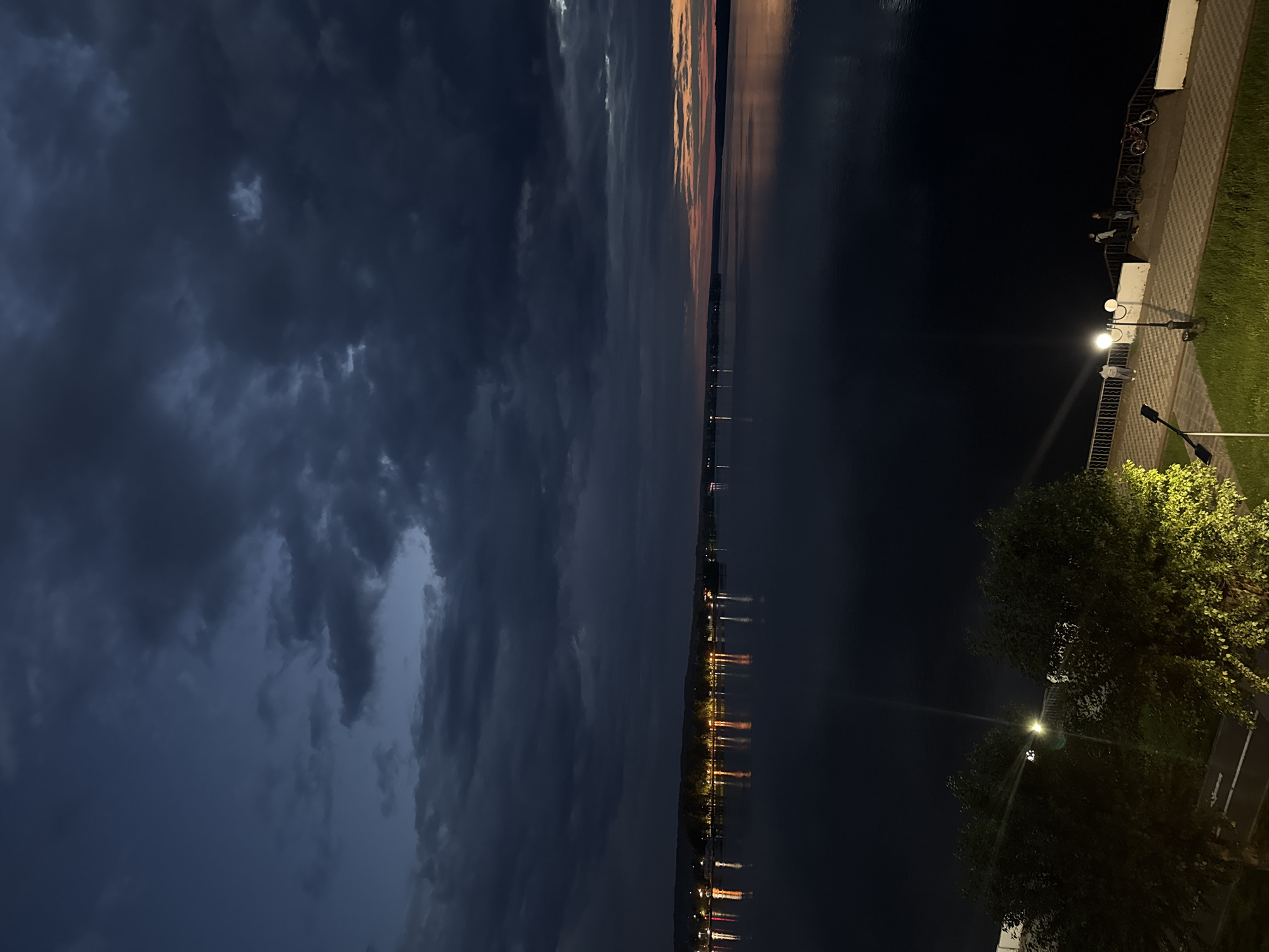 The image captures a dusky view of the Votkinsk Reservoir in Votkinsk, Russia. Dark, moody clouds dominate the upper portion of the frame, hinting at an approaching storm or the end of a rainy day. Below the dramatic sky, a wide expanse of dark water stretches towards a distant shoreline. Lights from a town or settlement are visible along the horizon, their reflections shimmering on the water's surface. In the foreground, a paved walkway runs alongside the reservoir. Several streetlights illuminate sections of the walkway, casting pools of light on the patterned pavement. A few individuals are present on the walkway, some standing near the railing and observing the water. Two bicycles are parked on the walkway near a group of people. Lush green grass borders the walkway, and trees with illuminated foliage are visible on the left side of the frame. The overall atmosphere is serene, with a touch of dramatic natural beauty from the turbulent sky and calm water.