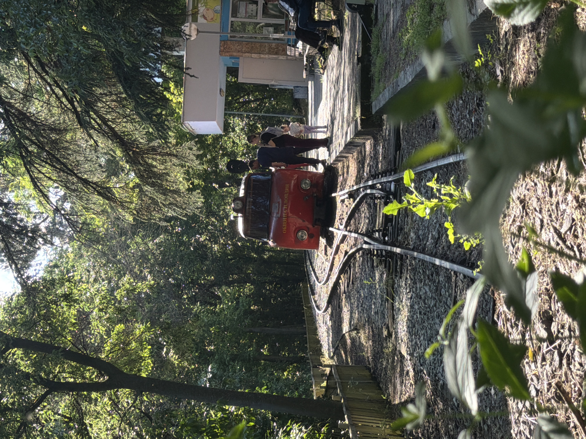 The image shows a narrow-gauge railway in Plovdiv, Bulgaria. A red locomotive is on the tracks, and several people are nearby. In the background is a train station building. The area is surrounded by trees and vegetation, creating a park-like setting. The lighting indicates it is likely daytime, with shadows cast on the tracks and surrounding foliage.