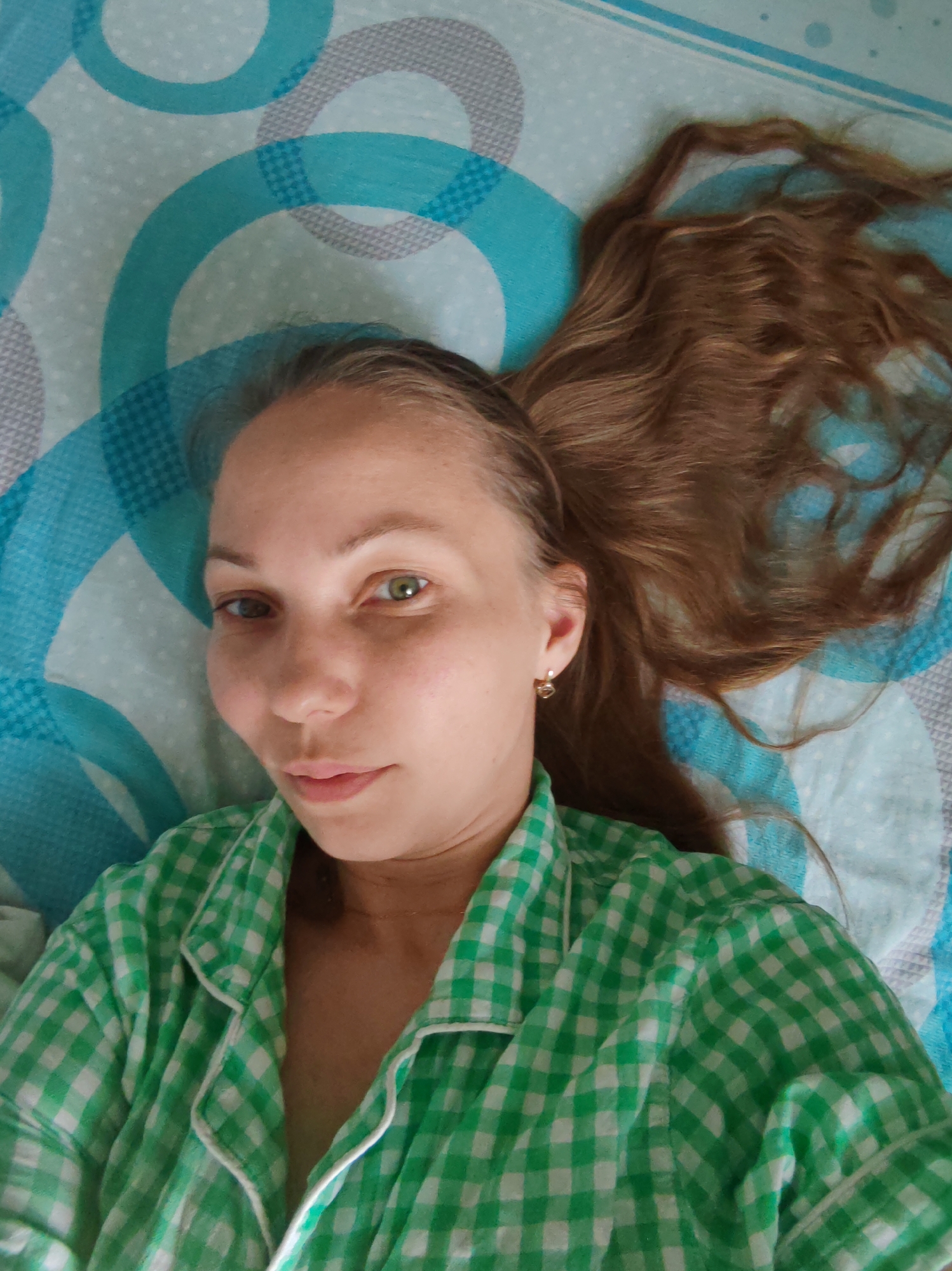 A light-skinned woman with brown hair is lying on a bed covered with a blue and gray patterned sheet. She is wearing a green and white checkered pajama top and a small gold earring. Her hair is spread out on the bed. The location is not evident in the image.