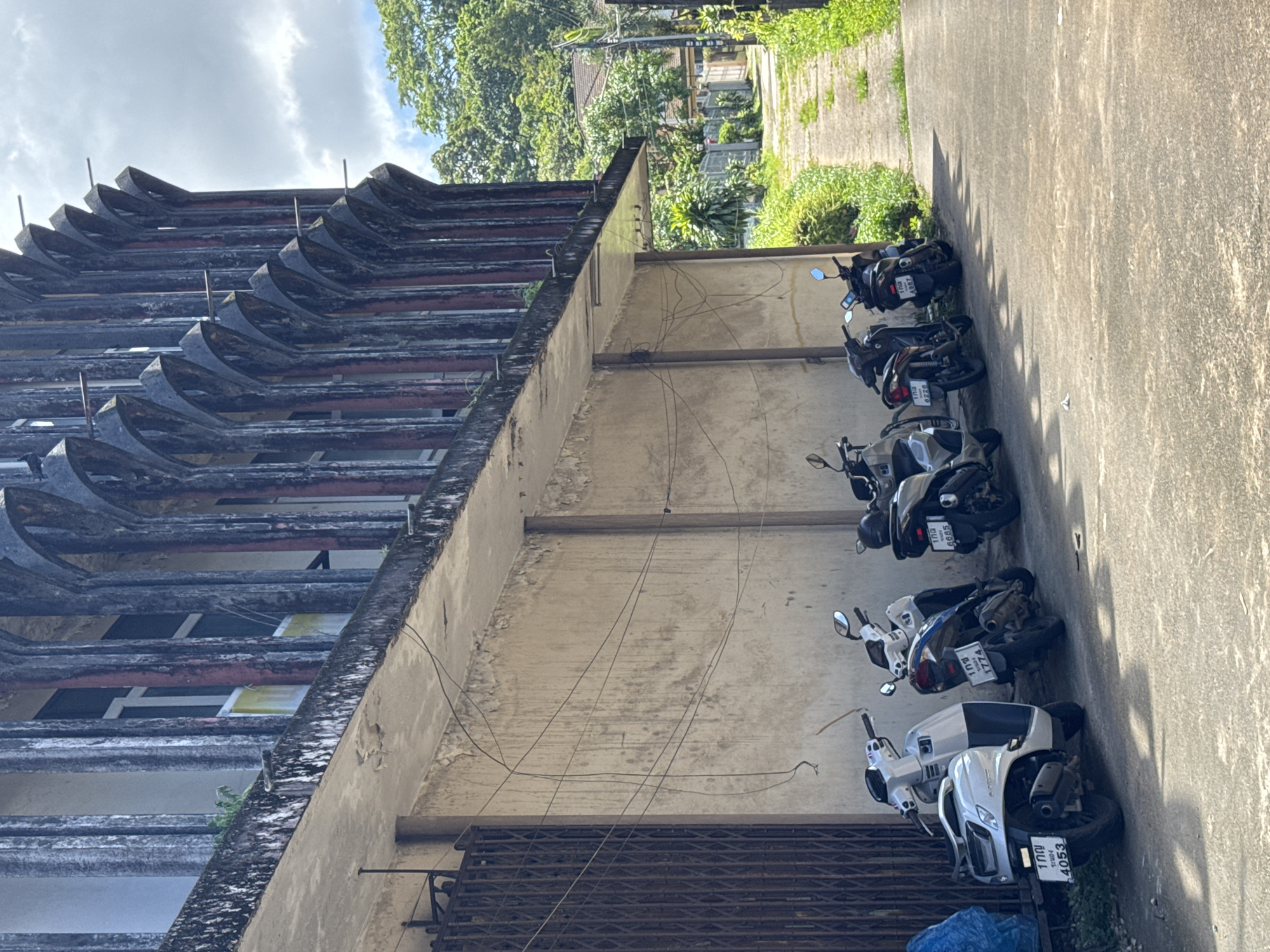 The image depicts the exterior of a weathered building in Ranong, Thailand, characterized by a distinctive facade of vertical, arched concrete structures. A row of parked motorcycles is visible in front of the building. The building's walls show signs of age and disrepair, with peeling paint and a network of overhead wires. To the right of the building, a narrow, unpaved pathway lined with lush green vegetation recedes into the distance, suggesting a residential or less developed area of the city. The sky is partly cloudy.