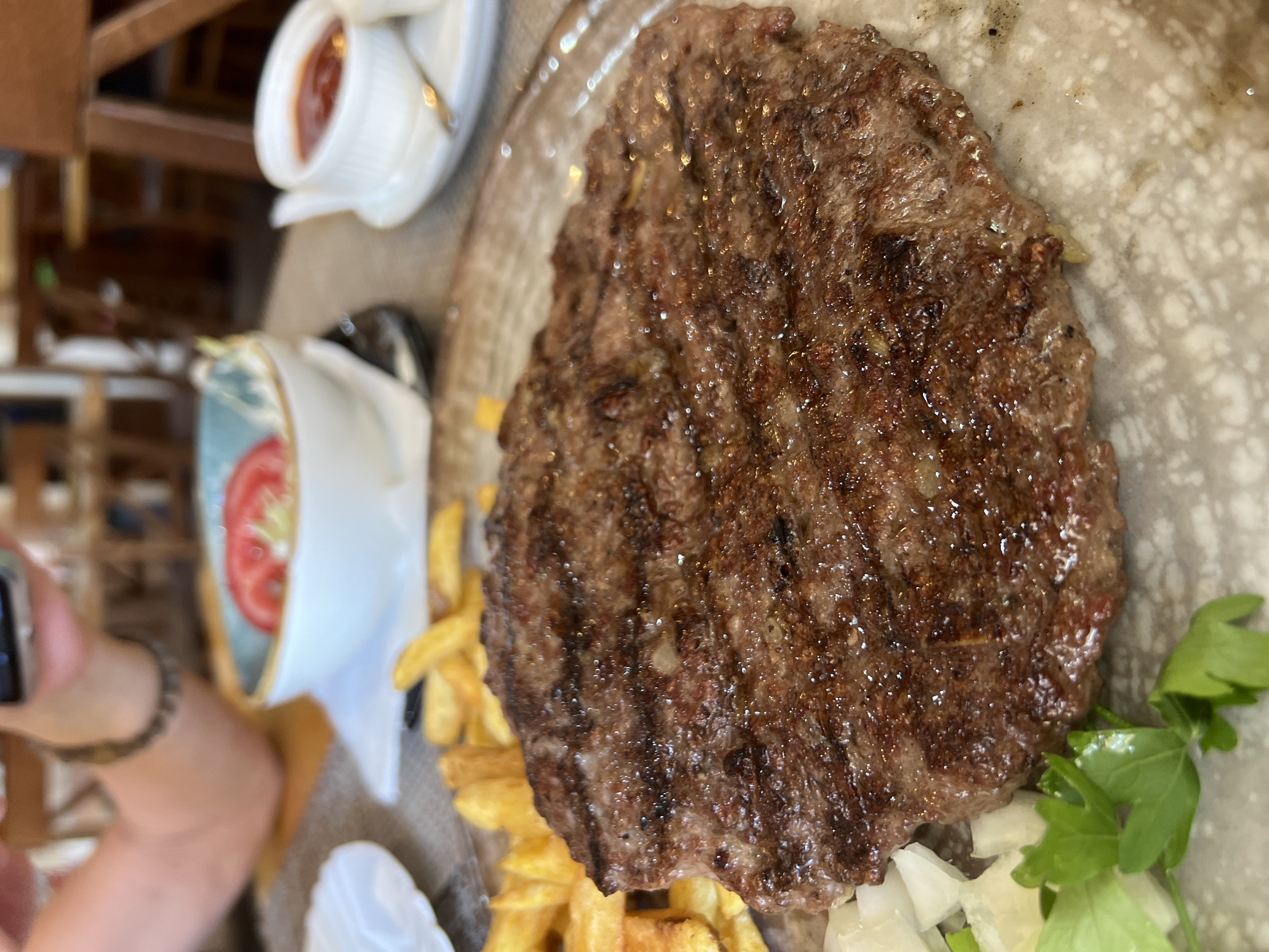 A close-up, overhead shot shows a grilled meat patty with grill marks, served on a speckled ceramic plate. French fries are piled behind the patty. To the left of the patty, chopped white onions and a sprig of parsley are visible. In the background, a bowl with sliced tomatoes and a small white ramekin with a red sauce are on a table. A person's hand holding a smartphone is visible in the upper left corner. The setting appears to be a restaurant in Budva, Montenegro.