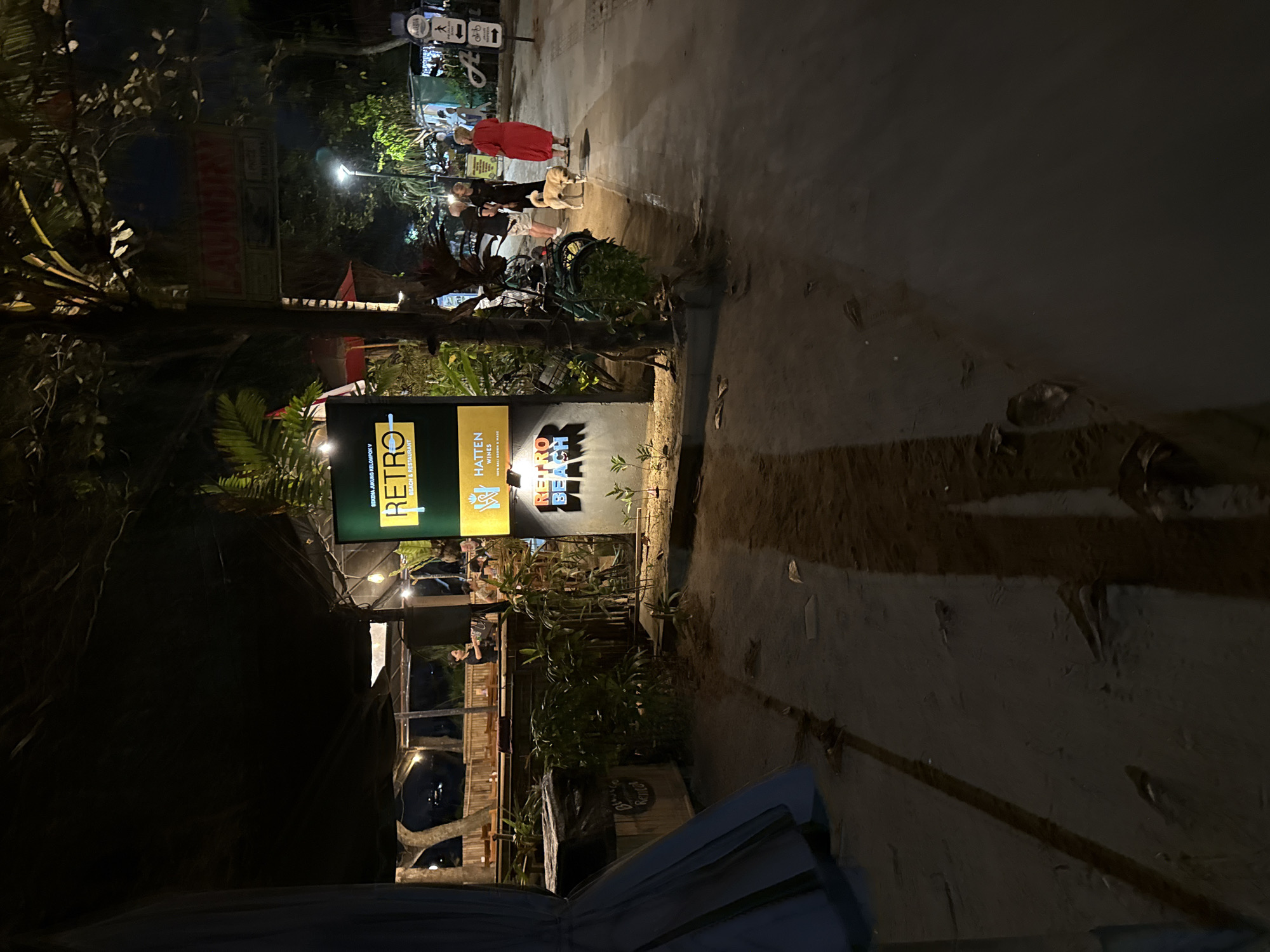 A nighttime exterior view shows a sign for "RETRO BEACH & RESTAURANT" in Denpasar, Indonesia. The sign is illuminated and features the word "RETRO" in large, yellow block letters with a horizontal yellow line above it. Below "RETRO," the words "BEACH & RESTAURANT" are written in a smaller font. A smaller sign below says "HATTEN WINES." The area appears to be a beachside establishment with sand on the ground and tropical foliage. In the background, there are people walking and a dog. A structure with tables and chairs, possibly part of the restaurant, is visible on the left. Another sign in the distance reads "LAUNDRY." The overall scene depicts a casual, lively evening setting near the coast.