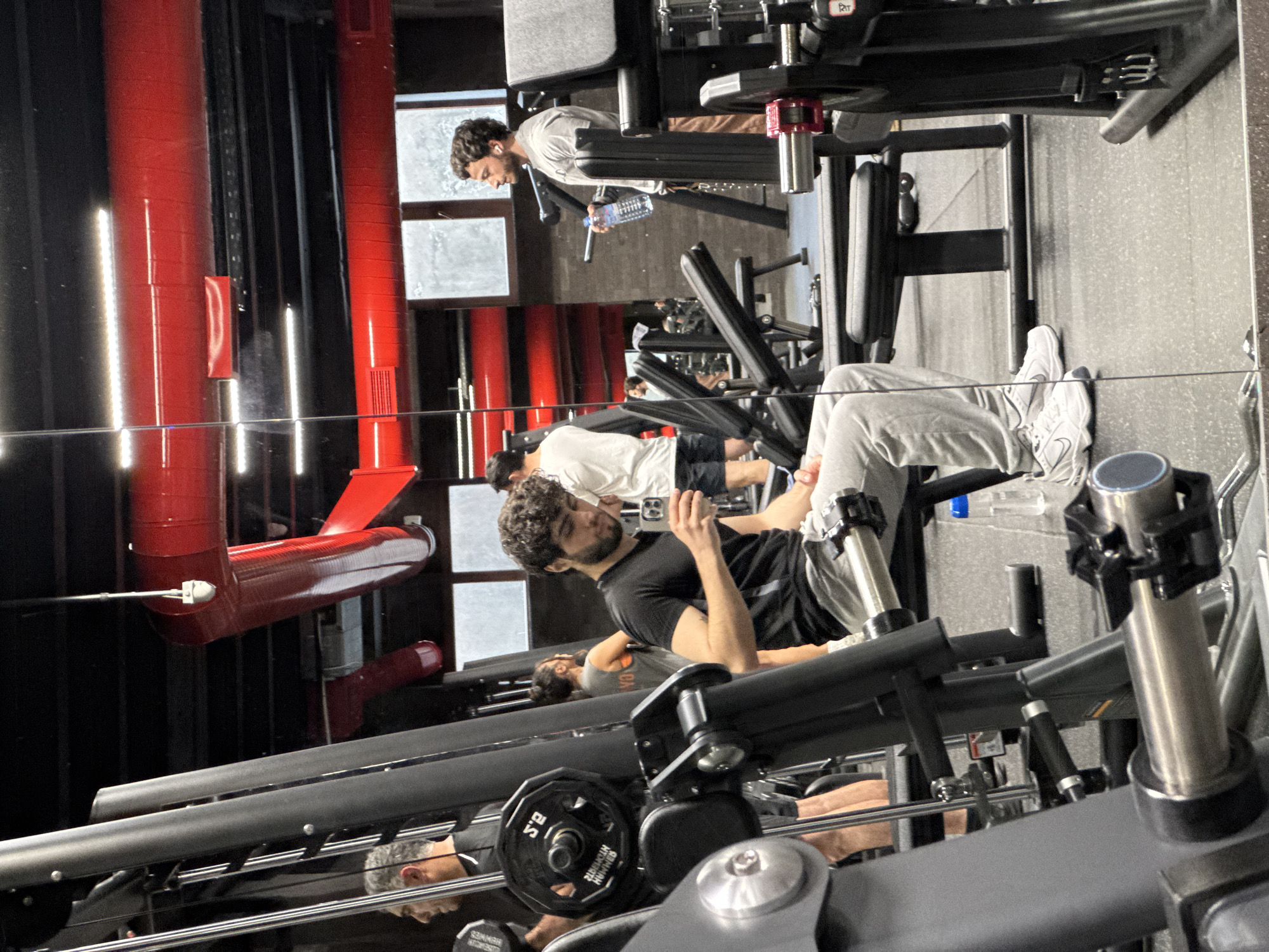 The image captures a scene inside a gym, likely located in Yerevan, Armenia, given the context provided. Several individuals are engaged in exercise routines, utilizing various gym equipment.
A man with curly hair, wearing a black shirt and grey sweatpants, is seated on a leg press machine, using his phone to take a picture. His feet are clad in white sneakers. Behind him, another individual, wearing a white shirt and black shorts, is positioned on another machine, possibly a leg extension or hamstring curl.
In the background, another man, also in workout attire, is seen holding a water bottle and appearing to be taking a break.
The gym itself features modern equipment and a unique aesthetic, with red ductwork contrasting against the dark-colored walls and equipment. The lighting is bright, and the atmosphere seems energetic.
The gym equipment includes leg press machines with weight plates. A reflective surface or mirror is present, showing the reflections of the individuals and equipment.