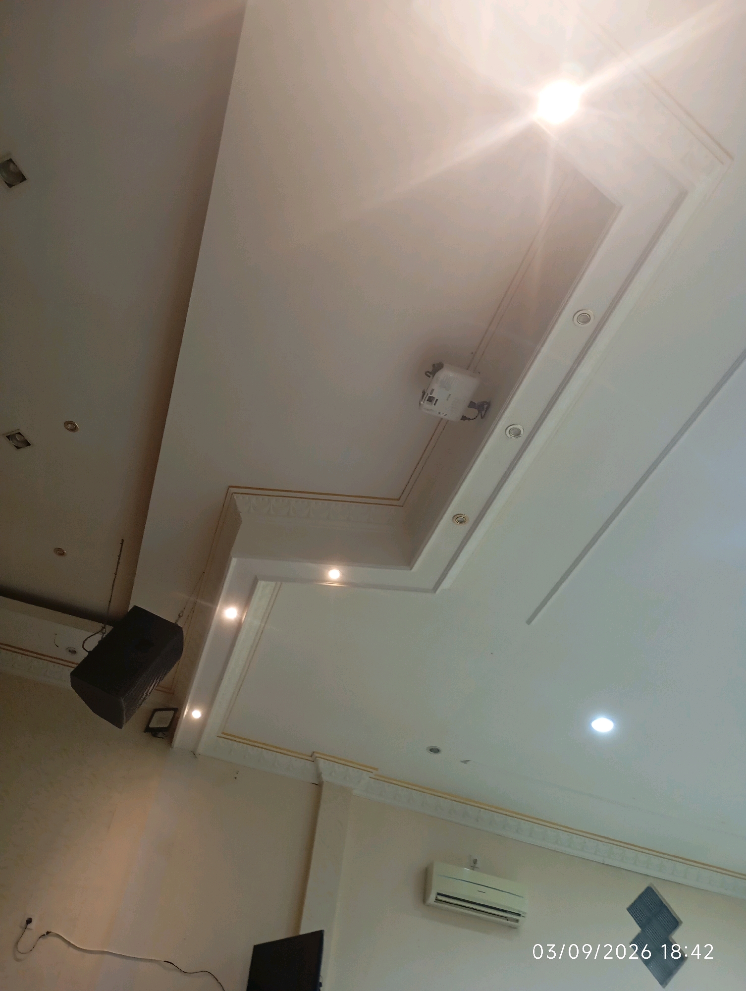 The image depicts the interior of what appears to be a meeting room or event space, likely in Bangunsari, Indonesia. The ceiling is a prominent feature, with a tiered design and embedded lighting fixtures. A projector is mounted on the ceiling. A black speaker is suspended from the ceiling on the left side of the frame. An air conditioning unit is visible on the wall on the right. The walls are a neutral color, with decorative molding along the top. A portion of a black television screen is visible in the bottom right corner. A timestamp "03/09/2026 18:42" is overlaid on the image. There are no people visible in the frame.