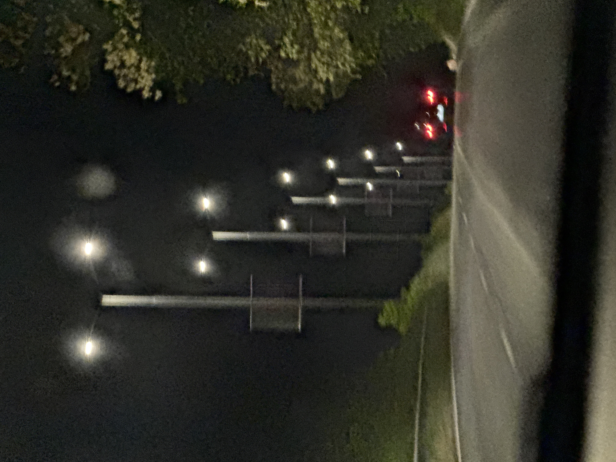 At night in Ledenik, Bulgaria, a roadway with multiple overhead streetlights and highway signs leads towards the horizon. The taillights of a vehicle are visible in the distance. Trees line the roadside on one side. A large, dark object is present in the foreground on the right, partially obstructing the view.