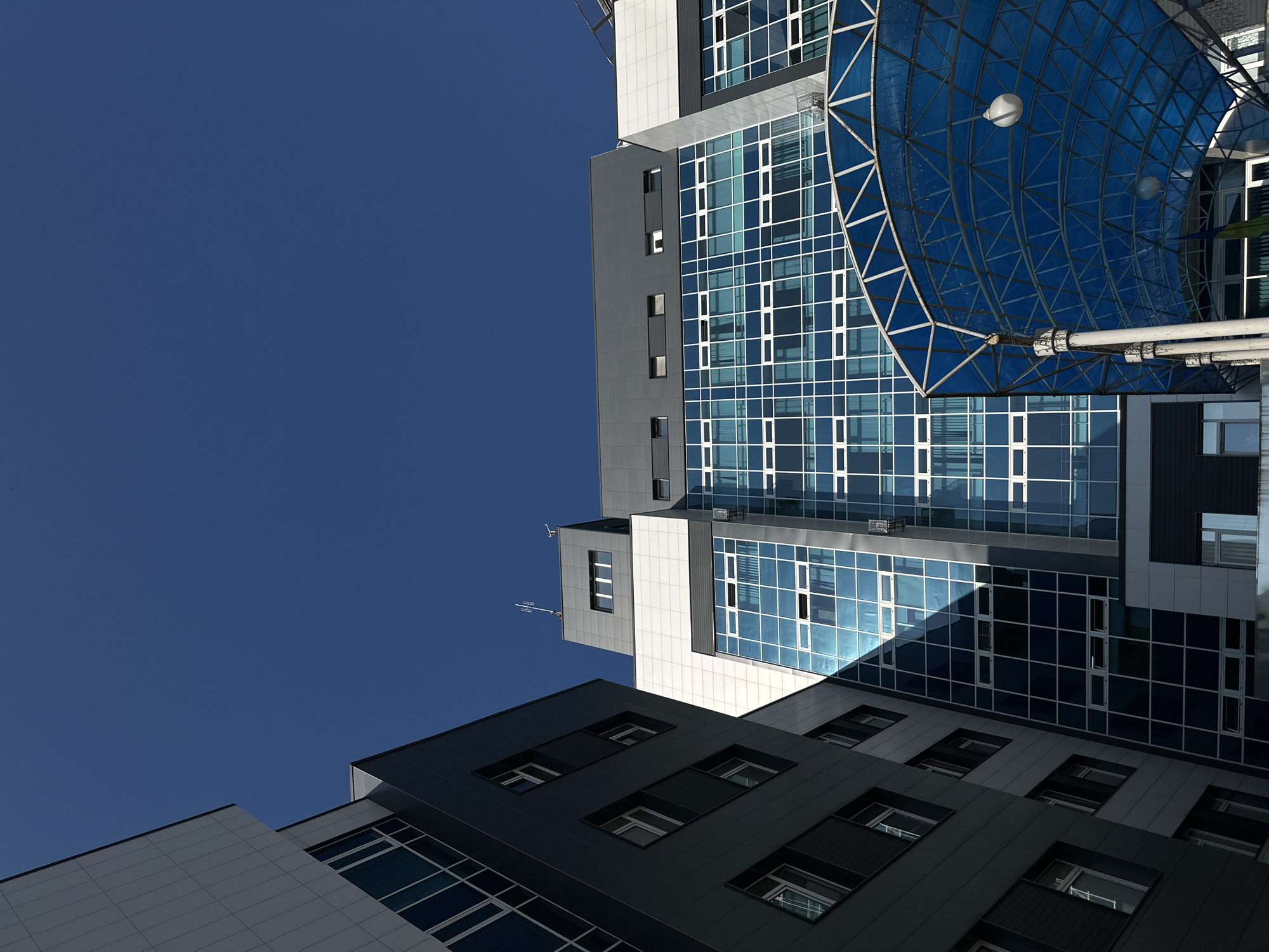 This is a low-angle, full-shot, vertical photograph of modern buildings under a clear, deep blue sky in Kyiv, Ukraine. The buildings feature a mix of dark gray paneling and large expanses of blue-tinted glass windows with white frames. The architectural styles are contemporary and angular. In the foreground, a blue, semi-transparent canopy with a white metal frame arches over an entrance. The sunlight casts sharp shadows, highlighting the geometric shapes of the structures. There are no people or discernible actions visible in the image. The scene conveys a sense of urban architecture and clear weather.