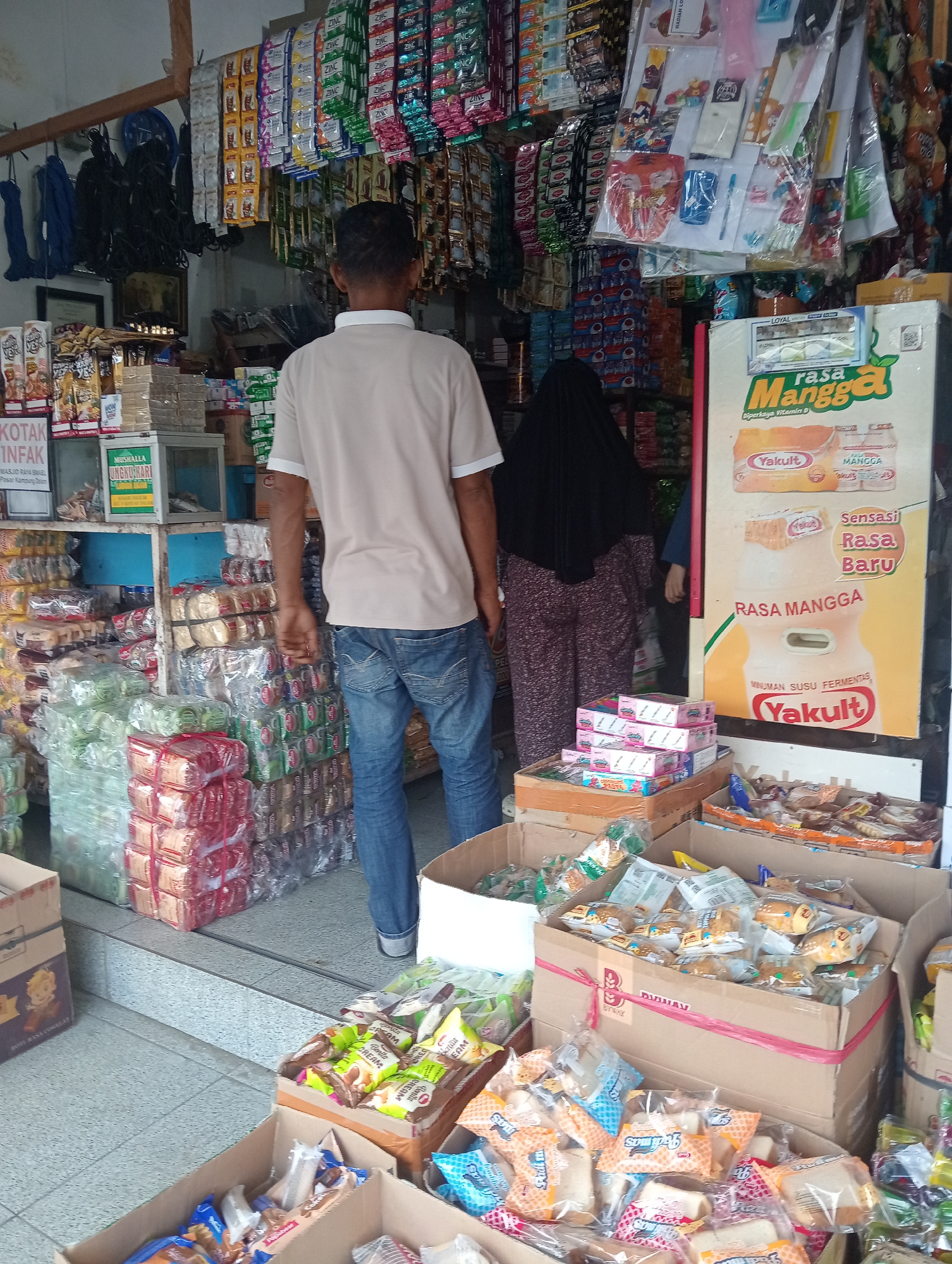 The image depicts the interior of a small shop in Pariaman, Indonesia, likely a convenience store or minimarket. In the foreground, several cardboard boxes are filled with packaged snacks, many of which appear to be biscuits or bread. A man wearing a light-colored t-shirt and blue jeans stands facing away from the camera, looking into the store. Behind him, a woman wearing a black hijab and patterned trousers is also present. The shop is densely stocked with various goods, including shelves lined with packaged food items, drinks, and hanging bags of candy or snacks. A large advertising poster for "Yakult Rasa Mangga" is prominently displayed on the right side of the image. The overall impression is that of a busy, local retail establishment with a wide array of products for sale.