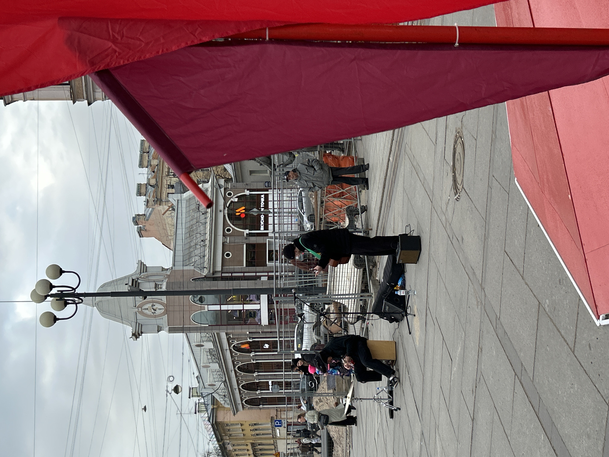 This image captures an outdoor street scene in Saint Petersburg, Russia, featuring a live music performance under an overcast sky. The foreground is partially obscured by large red and maroon fabric awnings or tarps.
In the center of the frame, two musicians are set up on a grey tiled sidewalk. One musician stands, dressed in dark clothing, appearing to play a stringed instrument like a guitar, with a microphone stand positioned nearby. The second musician is seated on a light brown cajon drum, also in dark attire. Various pieces of musical equipment, including speakers and a cymbal stand, are arranged around them.
Behind the performers, a small group of people are gathered, observing the performance. Metal barriers separate the performance area from what appears to be a public street or further pedestrian zone, where a silver car is partially visible.
The background is dominated by classical European architecture. One notable building features a prominent clock face and a dome-like structure on its roof. Another building exhibits arched windows and decorative elements. On a building facade, the Cyrillic text "ВКУСНО - И ТОЧКА" (VKUSNO - I TOCHKA), meaning "Delicious - Full Stop," is clearly visible, identifying a fast-food establishment. Numerous utility wires crisscross the cloudy sky above. A multi-globe lamppost stands to the left, and a blue "P" sign, indicating parking, is also visible. The scene depicts a lively daytime urban environment.