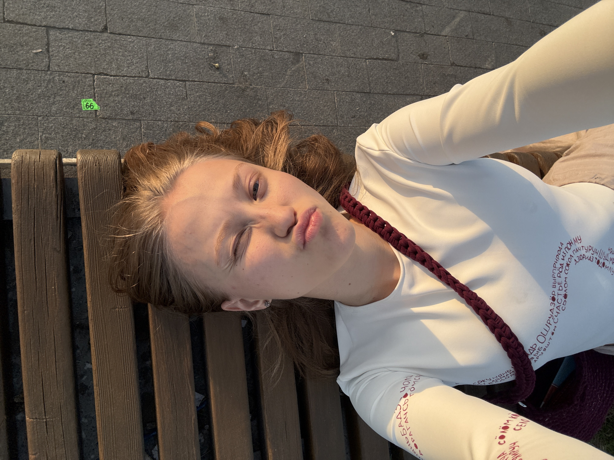 The image shows a young woman lying on a park bench. She is wearing a white long-sleeved shirt with circular text designs on the sleeve and chest and has a braided strap across her chest. She is making a pouting face and winking. The bench is wooden, and the ground nearby is paved with gray stone bricks. A green sticker with the number "66" is affixed to the ground. Given the OCR text on the shirt, it's likely the image was taken in the city of Perm, Russia.