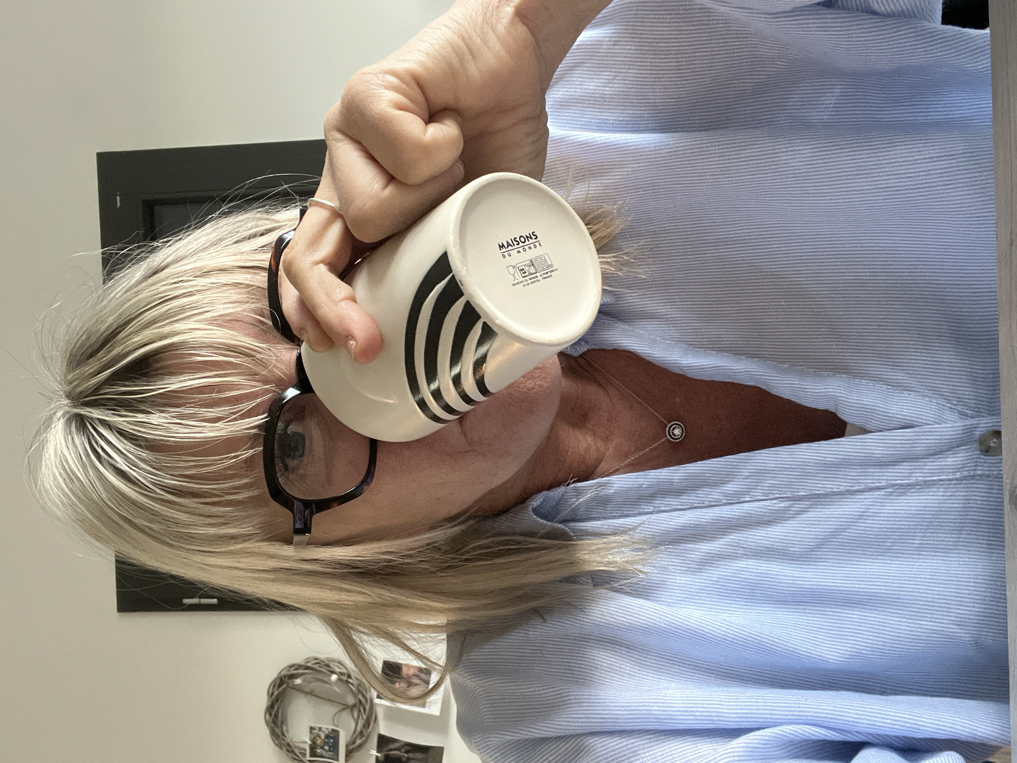 A middle-aged woman is drinking from a white mug with black stripes, with the bottom of the mug visible. The mug has the brand name "MAISONS DU MONDE". She is wearing glasses and a light blue pinstripe shirt with a silver necklace. There is a dark rectangular object on the wall behind her, and a decorative wreath with photos is visible in the lower-left corner. The city of Chambœuf, France, is not discernible from the image.