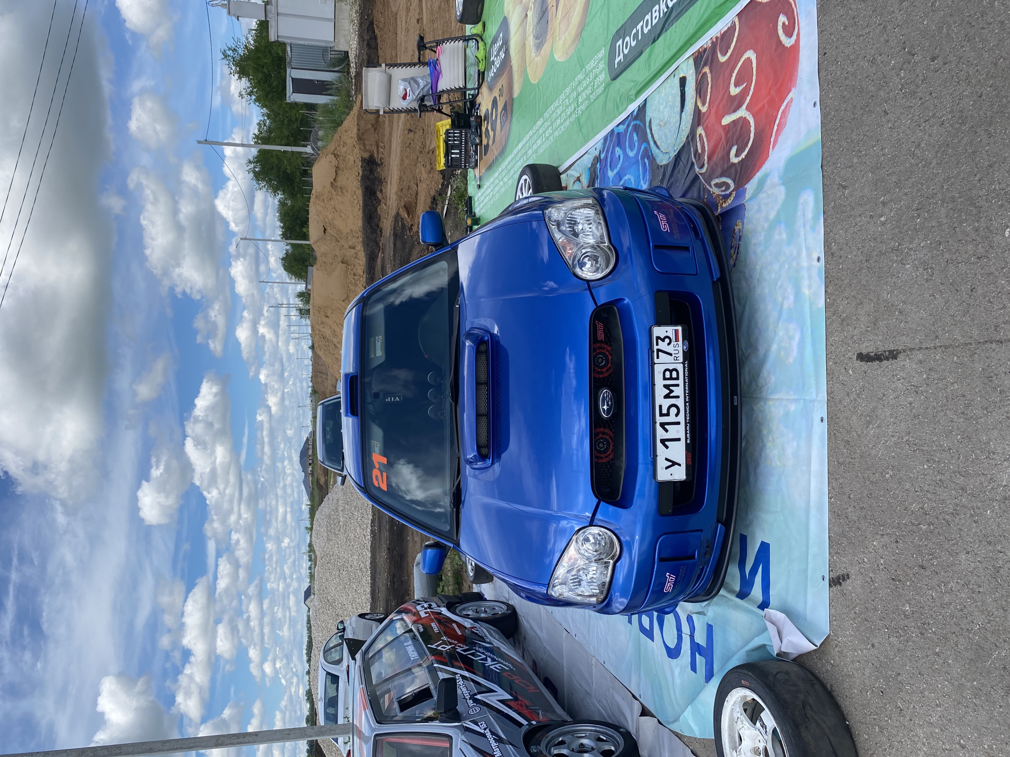 A blue Subaru WRX STI is parked on a white tarp with blue lettering on it, in what appears to be an outdoor event or race track setting. The car is facing the camera and has a bright orange number "21" on its windshield. To the left of the Subaru, another car with a black, white, and red livery is partially visible. In the background, there are piles of gravel and sand, suggesting a construction site or an area used for off-road activities. There are also buildings and trees in the distance. The sky is blue with scattered clouds. The location is specified as Svetloe Pole, Russia. A folded lawn chair and a toolbox are placed on a green tarp behind the Subaru.