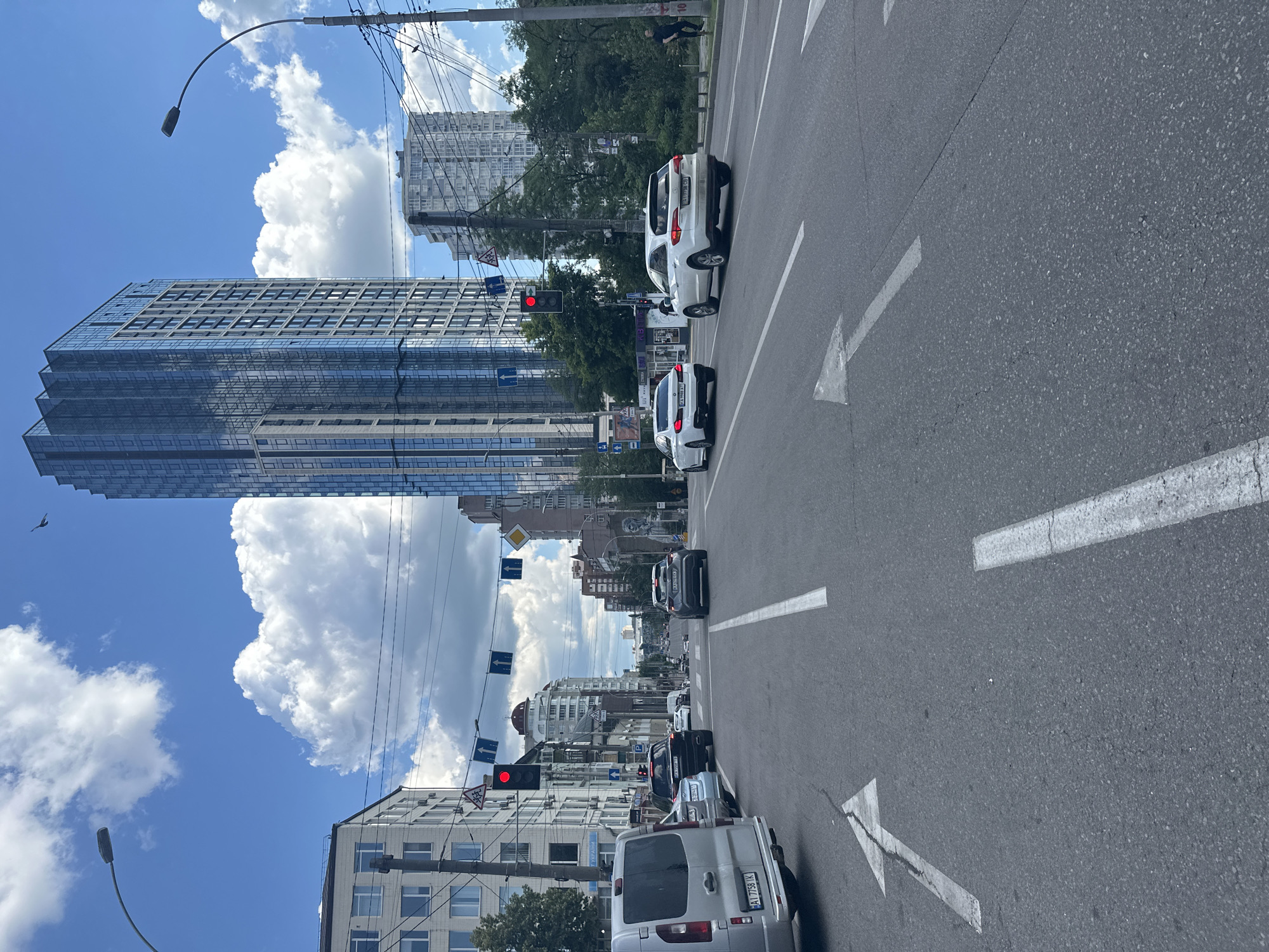 The image shows a street scene in Kyiv, Ukraine. The perspective is from the road, looking forward towards traffic. Several cars are visible, including a white SUV and a silver minivan. The road is paved and has white lane markings, including directional arrows. In the background, tall buildings line the street, some with modern glass facades and others with more traditional architecture. Trees are also present, suggesting a city park or boulevard.
The sky is blue with fluffy white clouds, indicating it is daytime and the weather is clear. A street lamp is visible on the left side of the frame. Traffic lights are also present, with one showing a red light, suggesting the cars are stopped or slowing down. The overall impression is a busy urban environment on a pleasant day. No people are clearly identifiable. The image is a still photograph.