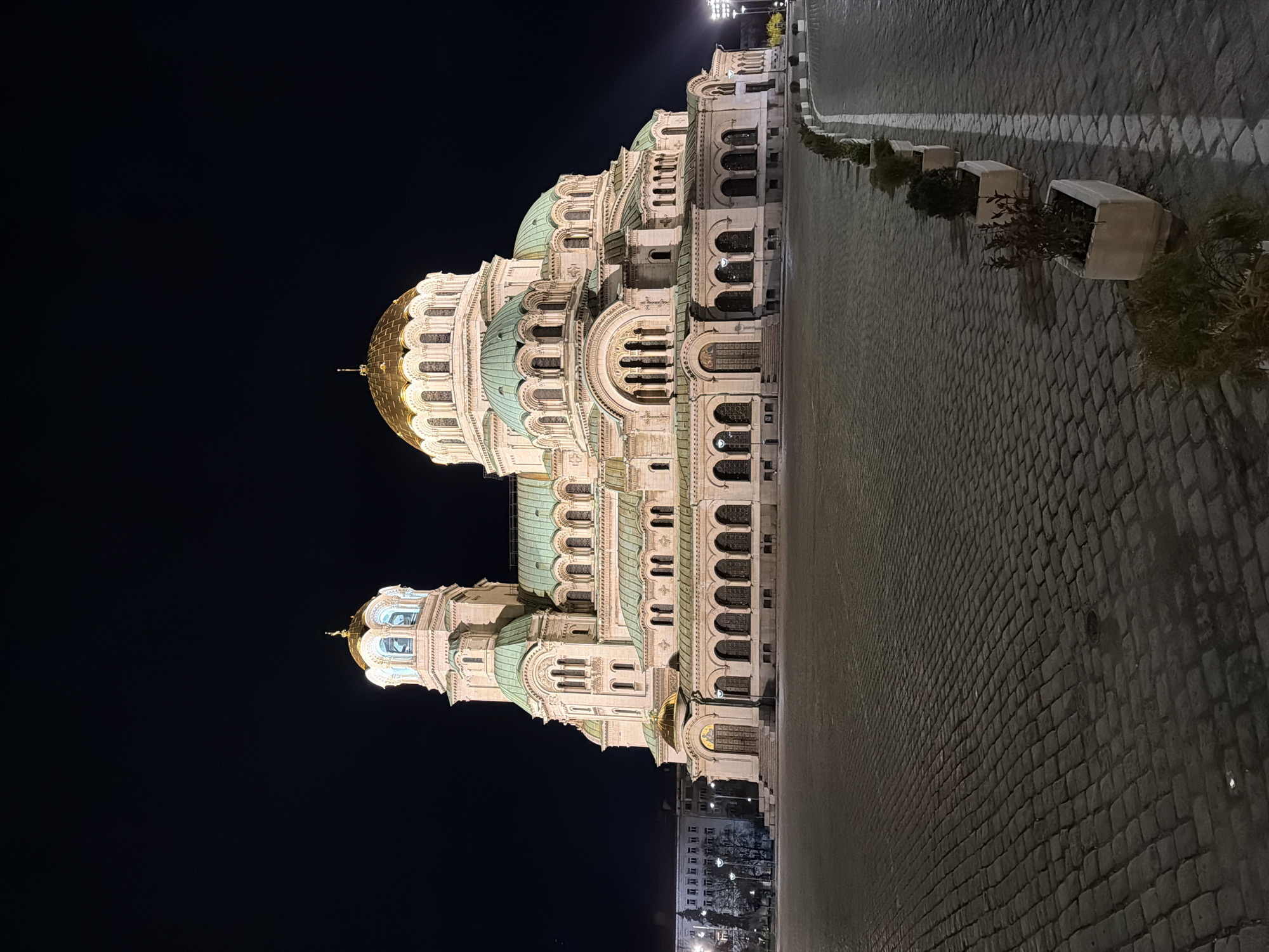 At night, the Alexander Nevsky Cathedral dominates the scene in Sofia, Bulgaria. The cathedral, illuminated, stands against a dark sky. To the side of the cathedral, a paved area stretches out, lined with flower pots. The paved area, reflecting some light, appears grey. Beyond this open area, other buildings and streetlights are visible.