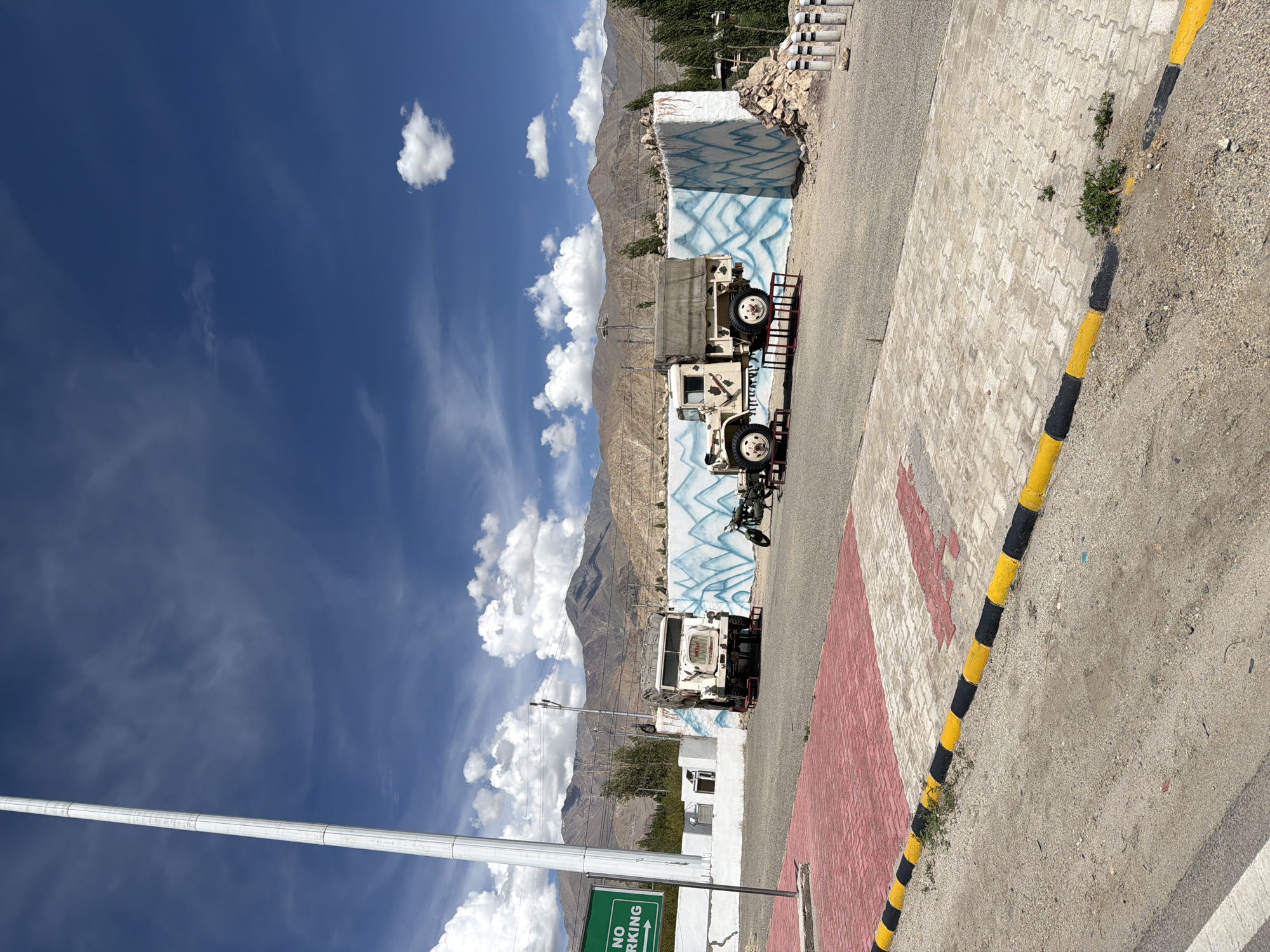 The image depicts a scene in the city of Leh, India, under a bright blue sky with scattered clouds. In the foreground, a paved area with red and white brick patterns is bordered by a yellow and black striped curb. Beyond this, a dusty, gravelly ground is visible, with a road running horizontally across the frame.
The central focus of the image is on two vintage military trucks displayed in front of a white wall adorned with abstract blue designs, possibly representing ice formations. The truck on the left is a white, boxy vehicle with a prominent grille. The truck on the right is a lighter-colored, open-top vehicle with a canvas cover over its rear bed. Between the trucks, a towed artillery piece is positioned, suggesting a military museum or exhibition.
Behind the trucks, the landscape transitions to a background of rugged, mountainous terrain with brown and grey hues. Green trees are scattered throughout the middle ground. A tall, white pole, possibly a flagpole or a light pole, stands prominently on the left side of the image, near a green sign that reads "NO PARKING" with an arrow pointing to the right. The overall environment suggests a public space, possibly near a roadside attraction or memorial. No people are visible in the scene.