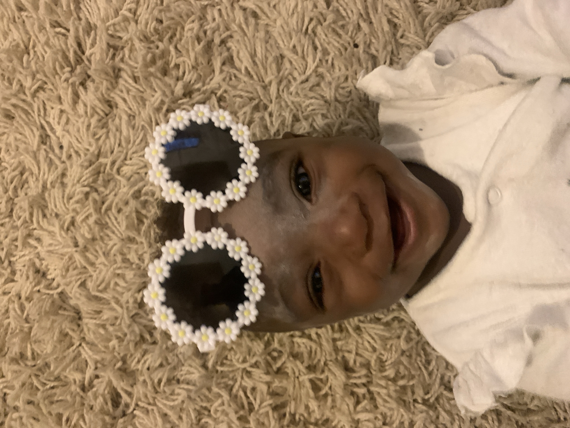 The image shows a baby lying on a shag carpet. The baby is wearing white clothes and daisy-themed sunglasses. The location is not discernible from the image.