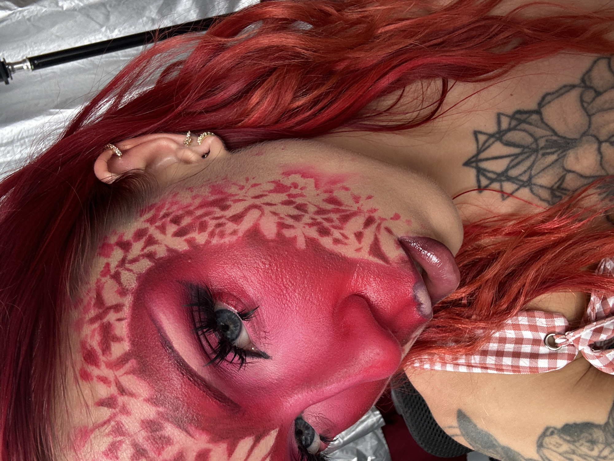 This media file features a close-up, low-angle shot of a person with bright red hair and elaborate pink and red face paint. The makeup is designed to resemble scales or intricate patterns around the eyes and across the forehead. The individual has dramatic eye makeup with long, dark eyelashes and a glistening, pink-toned lipstick. Their lips are slightly parted, with the tip of their tongue visible, also covered in pink lipstick.
Visible objects include several small, sparkling earrings on the visible ear, a tattoo on the neck and shoulder area with dark lines forming what appears to be a floral or geometric design, and a red and white checkered ribbon tied around the neck, possibly part of an outfit. The background is out of focus and appears to be a textured, possibly reflective surface in a light color, with a black rod-like object extending into the upper left corner.
The lighting is bright, highlighting the makeup and textures. The overall mood conveyed is artistic and perhaps a bit theatrical, focusing on the dramatic visual transformation through makeup. There is no visible text in the media file. The location context of North Kuta, Indonesia is provided externally and cannot be discerned from the image itself.