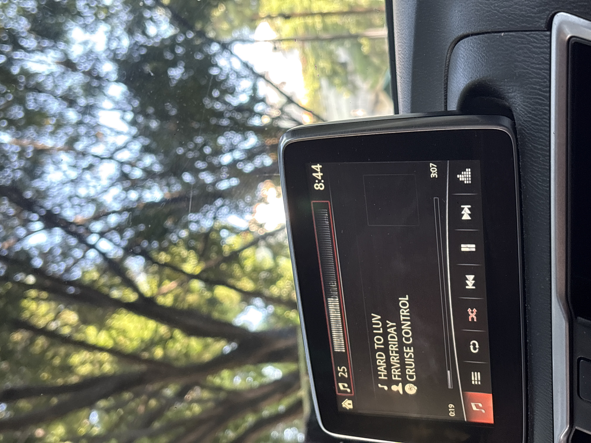 The image depicts the interior of a vehicle, with a display screen showing a music playback interface. The screen indicates a song titled "HARD TO LUV" by FRVRFRIDAY is playing. The time displayed is 8:44, and the song's progress is at 0:19 of a total 3:07. The volume is set at 25. Through the windshield, a blurry background of trees is visible. The location cannot be determined to be in Mexico City with the given information.