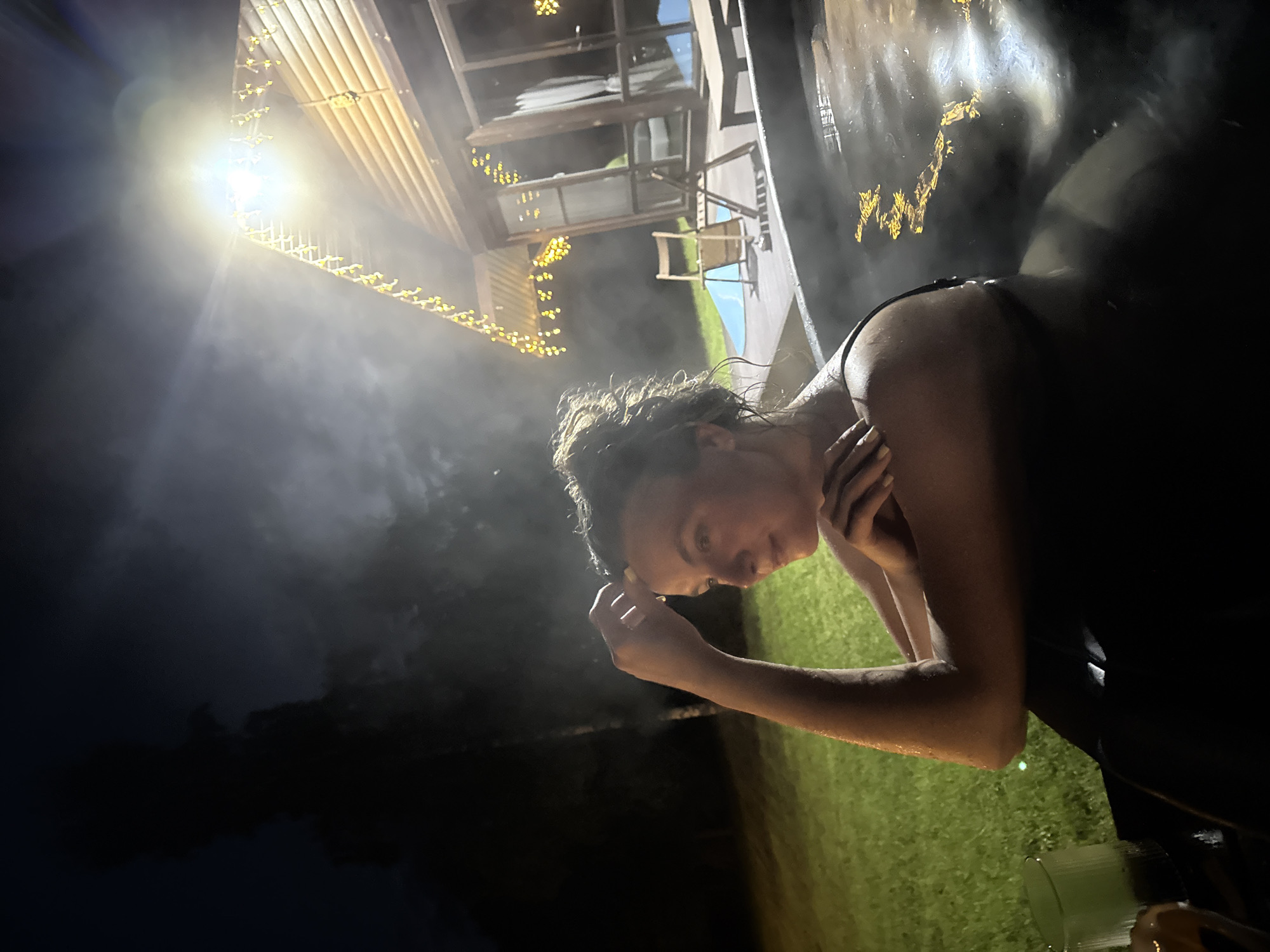 An outdoor night scene in Копнино, Russia, depicts a woman relaxing in a dark-colored hot tub, from which steam emanates. The woman, seen from the chest up, has wet, dark hair and rests her right hand near her forehead while looking towards the right side of the frame with a tranquil expression. She appears to be wearing a dark swimsuit. In the mid-ground, a brightly illuminated A-frame building, possibly a cabin or guesthouse, features numerous warm-toned string lights draped along its roofline and a powerful central light source that casts a significant glare and illuminates the surroundings. A wooden deck adjacent to the building holds a folded wooden chair, a small table, and a blue mat. To the left, a dark, grassy area with trees recedes into the background, dimly lit by the artificial light. In the bottom left foreground, a glass with liquid rests on a dark surface.