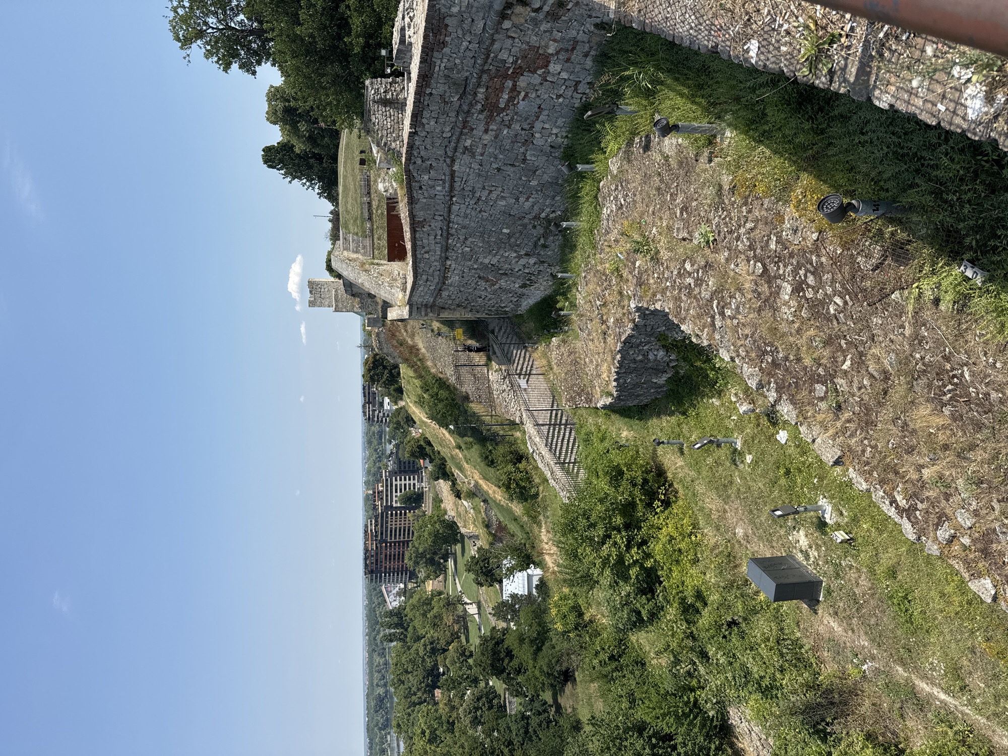 This is an eye-level, outdoor, medium shot of a historical fortification on a sunny day in Belgrade, Serbia. The camera is positioned looking down and to the right, overlooking a stone wall and a green, grassy slope dotted with trees. In the background, modern apartment buildings and a distant river are visible, suggesting an urban setting. The foreground shows a weathered stone pathway with several small, modern lights installed along its edge. A dark, rectangular electrical box is also visible on the slope. The sky is clear and blue, with a few wispy clouds. There are no people clearly visible in the image. The overall impression is one of historical ruins integrated into a modern city landscape.