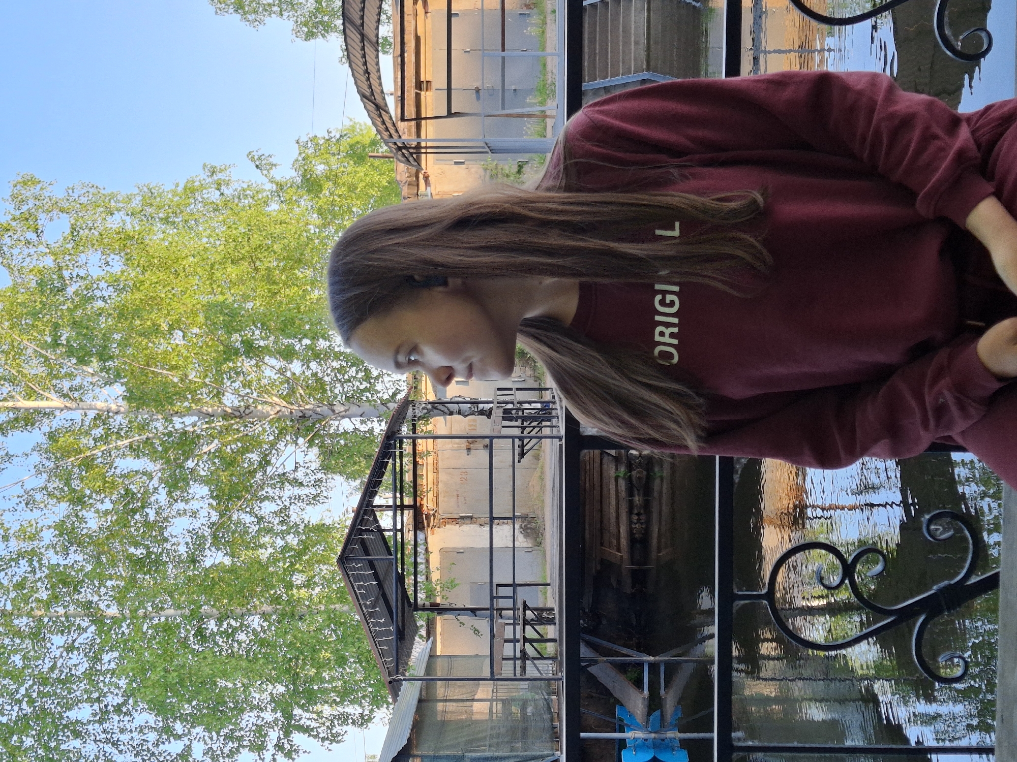 A young woman with long, light-brown hair wearing a burgundy sweatshirt with the word "ORIGINAL" on the front stands on an outdoor balcony with a black iron railing, overlooking a body of water. The water reflects the light from surrounding buildings and trees. The background features trees, buildings, and a sky with blue portions. The scene is likely located in or near Chaikovsky, Russia.