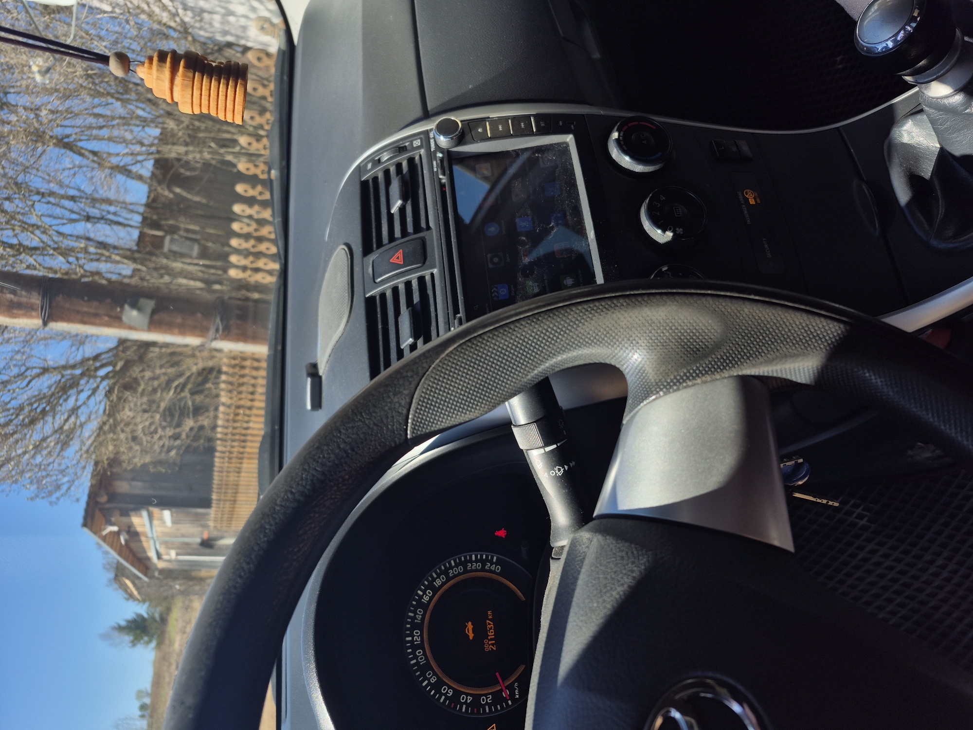 The image depicts the interior of a car, focusing on the dashboard and steering wheel. The car's instrument panel shows a speed of around 20 km/h and a mileage of 211,637 km. Warning lights are visible. The steering wheel has a textured surface. The dashboard features a central console with air vents, a hazard light button, and a touch screen display. Climate control knobs are located to the right of the display. A gear shift lever is also visible. A wooden air freshener hangs from the rearview mirror. Outside the car, a barn or rural building is visible in the background, suggesting a location in a rural or semi-rural area. No people are visible in the frame. The setting appears to be Дойная, Russia.