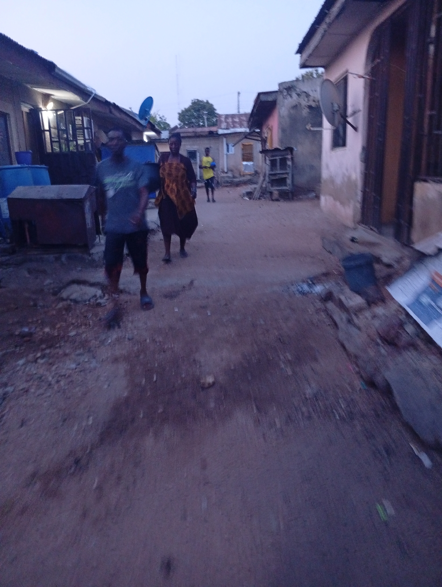 The photo captures a dirt road scene in Zango-ndaji, Nigeria. Three individuals are visible, with one person in a yellow shirt further down the road and two individuals closer to the camera. The closest individual is male, wearing a dark t-shirt and shorts. A woman in a patterned dress is walking beside him. The buildings are low-slung and some appear to have satellite dishes attached to them. The overall setting is a residential area with a rough road surface.