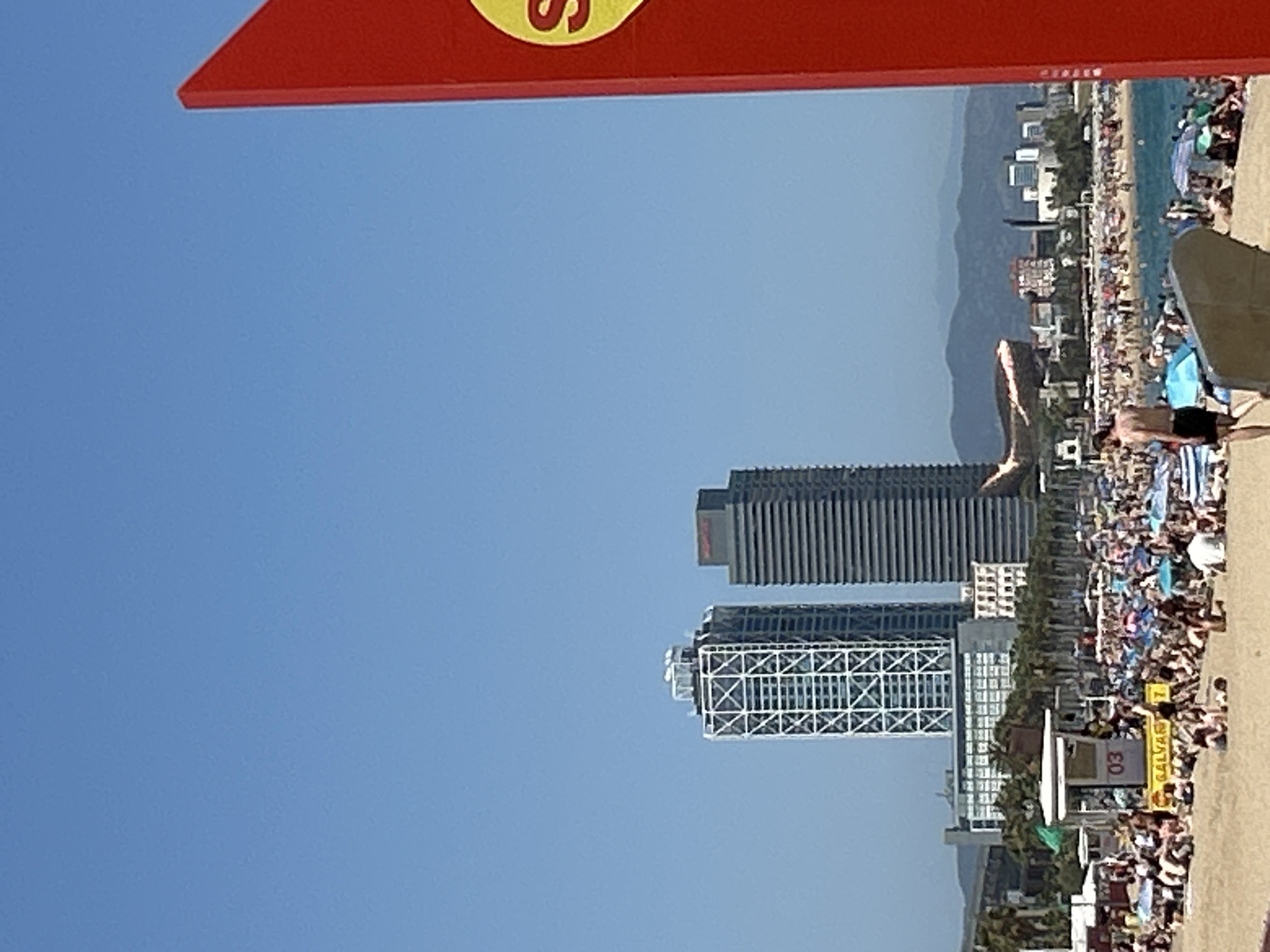 The image captures a vibrant beach scene in Barcelona, Spain, under a clear blue sky. In the background, two prominent skyscrapers stand tall, one with a distinct white geometric lattice facade and the other a sleek, dark rectangular structure. A low-rise building with many windows is visible between them, and further in the distance, a city skyline with more buildings and mountains can be seen. The foreground is dominated by a crowded beach filled with people relaxing under umbrellas and sunbathing on the sand. A lifeguard station, identified by the "03" and "GALVANI" signage, is situated on the beach. To the right of the frame, a large, bright red sign with a yellow circle and the letter "S" partially obscures the view. The sea is visible beyond the beach, with a scattering of boats on its surface. The overall atmosphere suggests a sunny and bustling day at the beach.