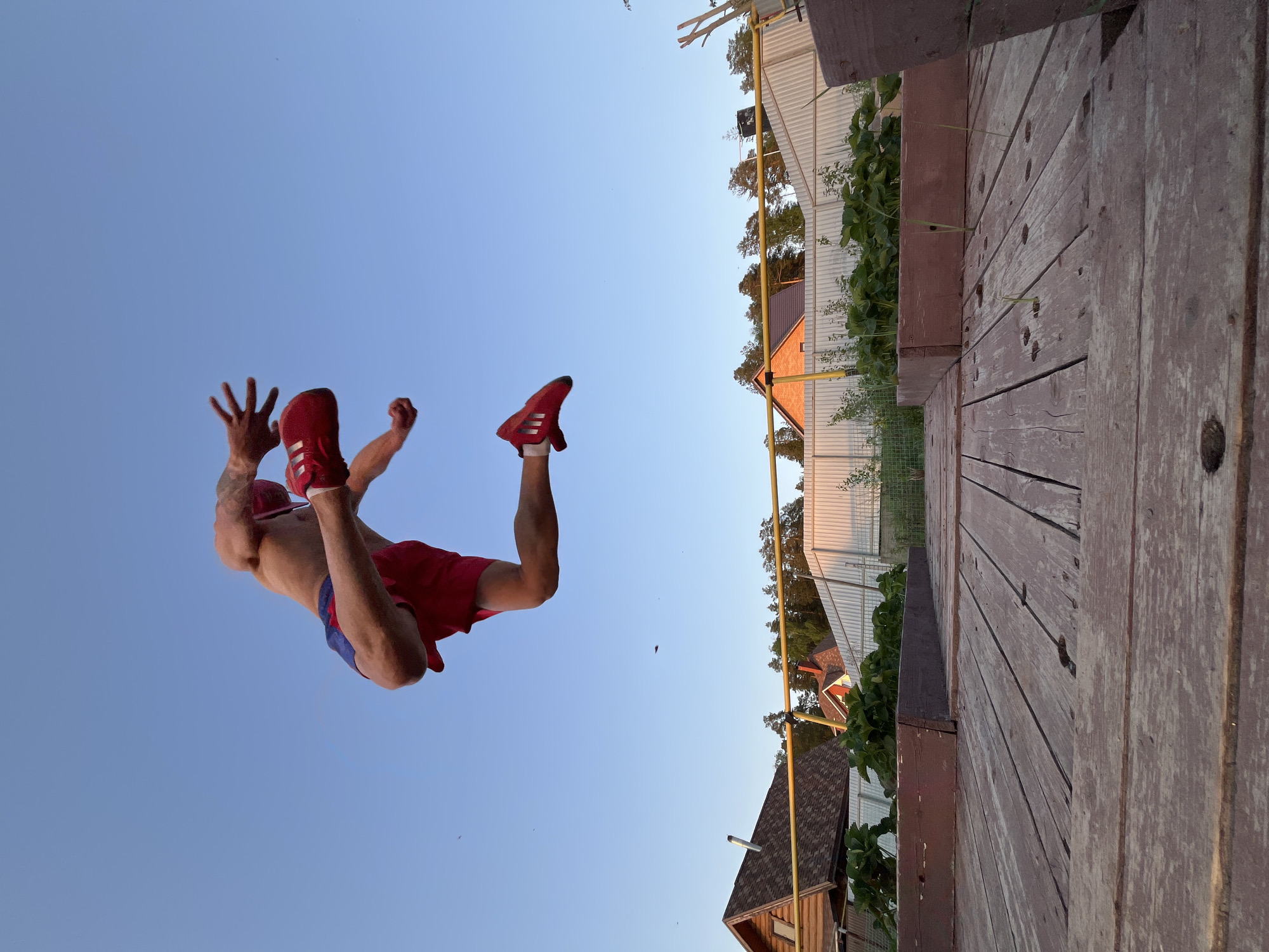 A man is in mid-air, performing a backflip in an outdoor setting. He is wearing red shorts, red Adidas shoes with white stripes, and a red hat or band. His torso is bare, and tattoos are visible on his left arm. The backdrop includes a clear blue sky. To the side, there's a wooden structure, likely a deck or elevated platform, with greenery visible nearby. In the distance, there are houses with varying roof styles, including gable and shed roofs, and a white fence. The image was taken in the city of Chaikovsky, Russia.