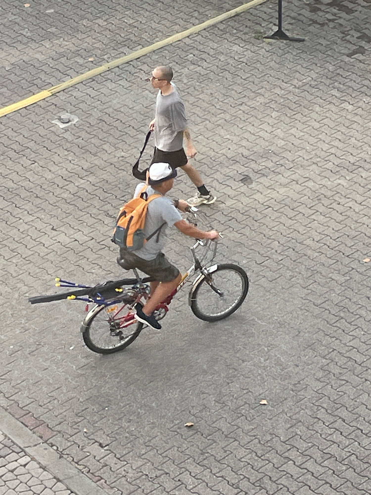 In Sochi, Russia, on a paved walkway constructed of interlocking rectangular blocks, two individuals are present. In the foreground, a person is riding a bicycle. This individual wears a white cap, a grey t-shirt, and camouflage-patterned shorts. An orange backpack is strapped to their back. The bicycle is red and has a black bag attached to the rear rack. Extending from the rear of the bicycle are several yellow-capped poles, possibly for fishing. Behind the cyclist, a second individual walks. This person has a shaved head, wears glasses, a grey t-shirt, and dark shorts. They are carrying a black bag slung over their shoulder. A yellow line is visible on the pavement to the left of the walkway. The overall environment appears to be outdoors, with sunlight casting shadows.