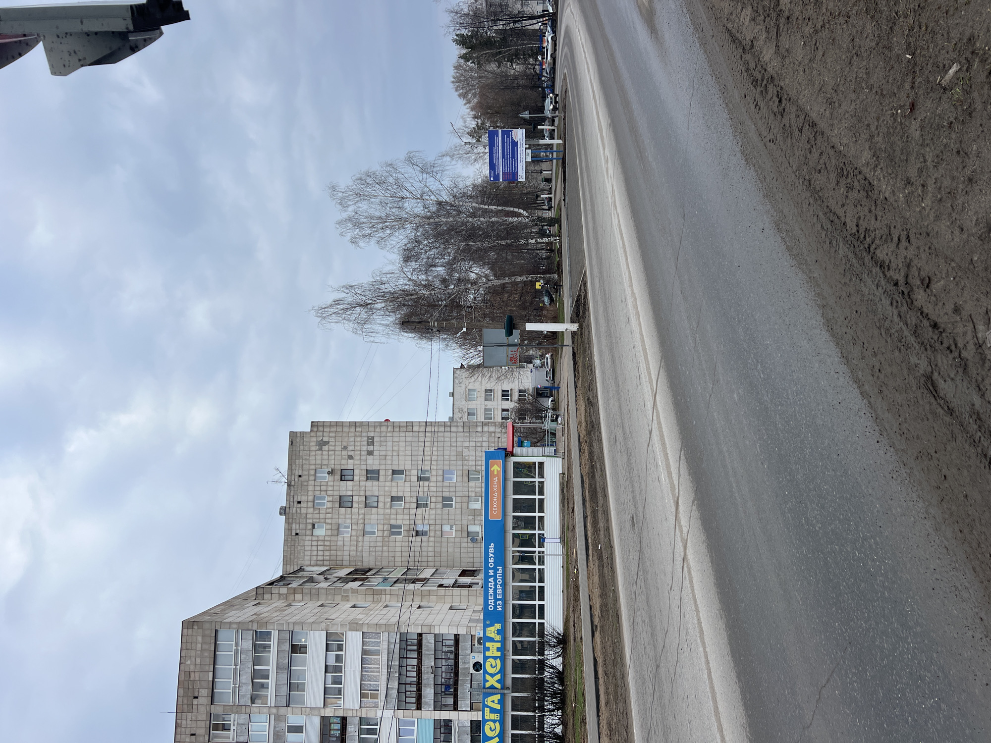 A street scene in Chaikovsky, Russia, presents a medium-wide, eye-level shot taken on a cloudy day. A grey, asphalt road stretches diagonally across the right half of the frame, showing some wear and tear. To the right of the road is a dirt shoulder.
The left side features a tall, Soviet-era apartment building, its facade composed of grey concrete panels and many windows. Attached to the base of the building is a one-story structure with a blue sign in Cyrillic that reads "SECONAD KHENDA."
Beyond the building, a row of leafless trees lines the roadside, followed by another building farther back. A blue and white sign sits next to the trees. Several vehicles are parked in the distance.