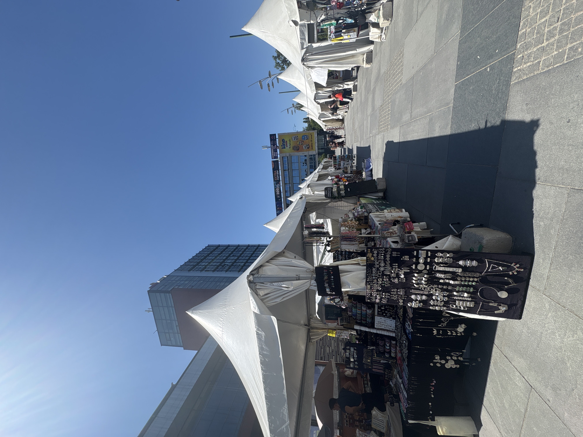 A street market is set up on a paved plaza with a clear blue sky above. White tents are erected in a row, displaying various goods such as jewelry, clothing, and other artisanal items. In the foreground, a tent displays an assortment of necklaces, earrings, and bracelets on a black velvet board. A person wearing a dark shirt is visible behind a display table on the left side of the image. Further down the plaza, other tents are set up, with a few individuals browsing the stalls. Buildings, including a modern high-rise with glass windows and a building with a large billboard, are visible in the background. The scene captures a bustling outdoor market environment.