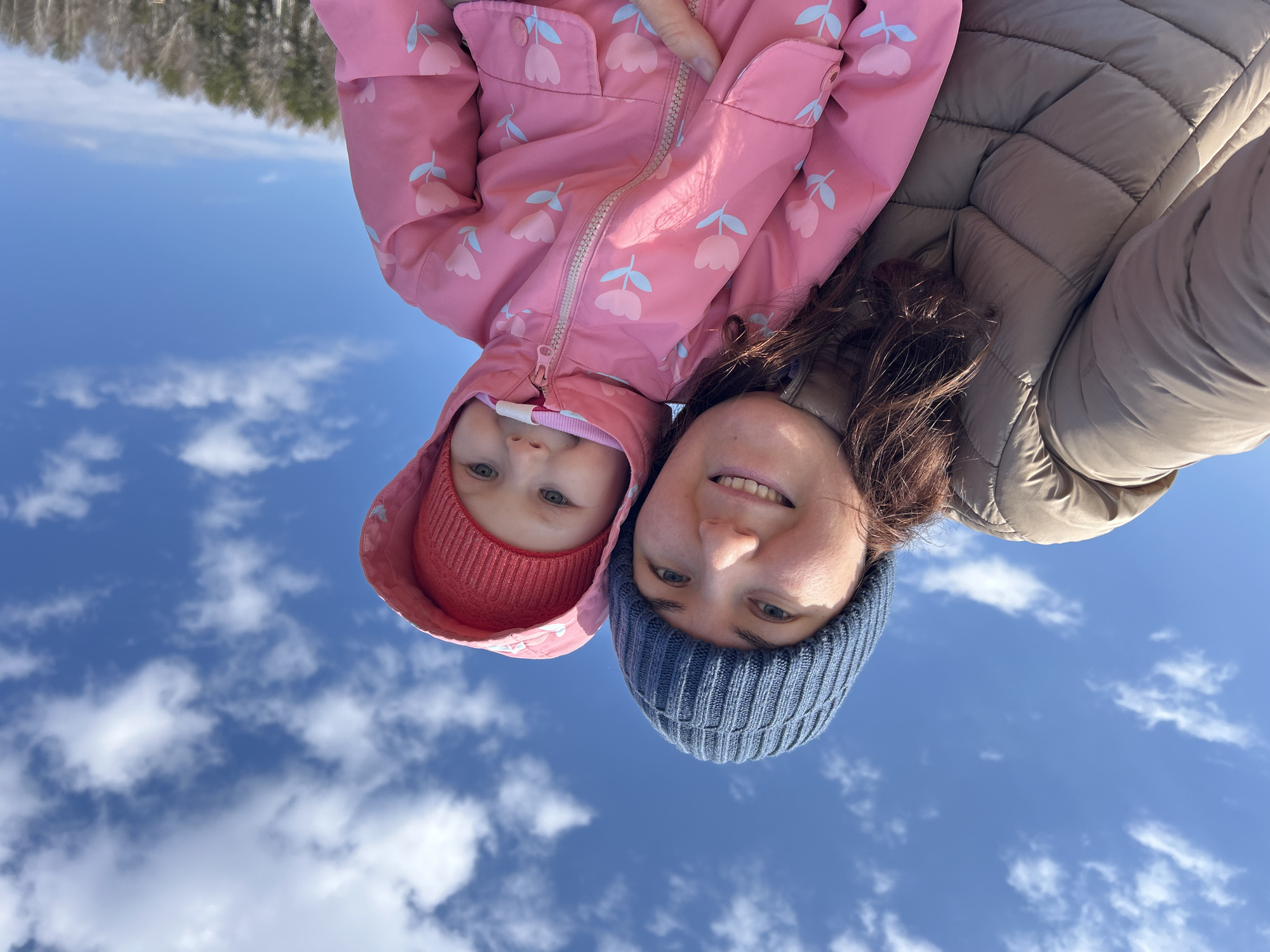 An upside-down selfie captures an adult and a young child, positioned against a bright blue sky filled with scattered white clouds. Their faces are at the lower part of the frame, smiling downwards at the camera.
The adult, on the right, wears a dark blue knitted beanie and a light brown or tan quilted puffer jacket. She has dark hair and a broad, joyful smile. The child, on the left, appears to be a toddler, sporting a red knitted hat under a pink hooded jacket. The pink jacket is adorned with a pattern of light blue, tulip-like flowers with green stems. The child looks at the camera with wide eyes and a slight, pleasant expression.
Both individuals are dressed in warm clothing, suggesting cool weather. In the upper portion of the image, visible above their bodies, is a blurred background of bare trees or a forest line, indicating an outdoor, natural setting. The vibrant blue sky and bright sunlight suggest it is daytime. The overall mood of the image is happy and intimate. No visible text is present. The photo was taken in Chaikovsky, Russia.