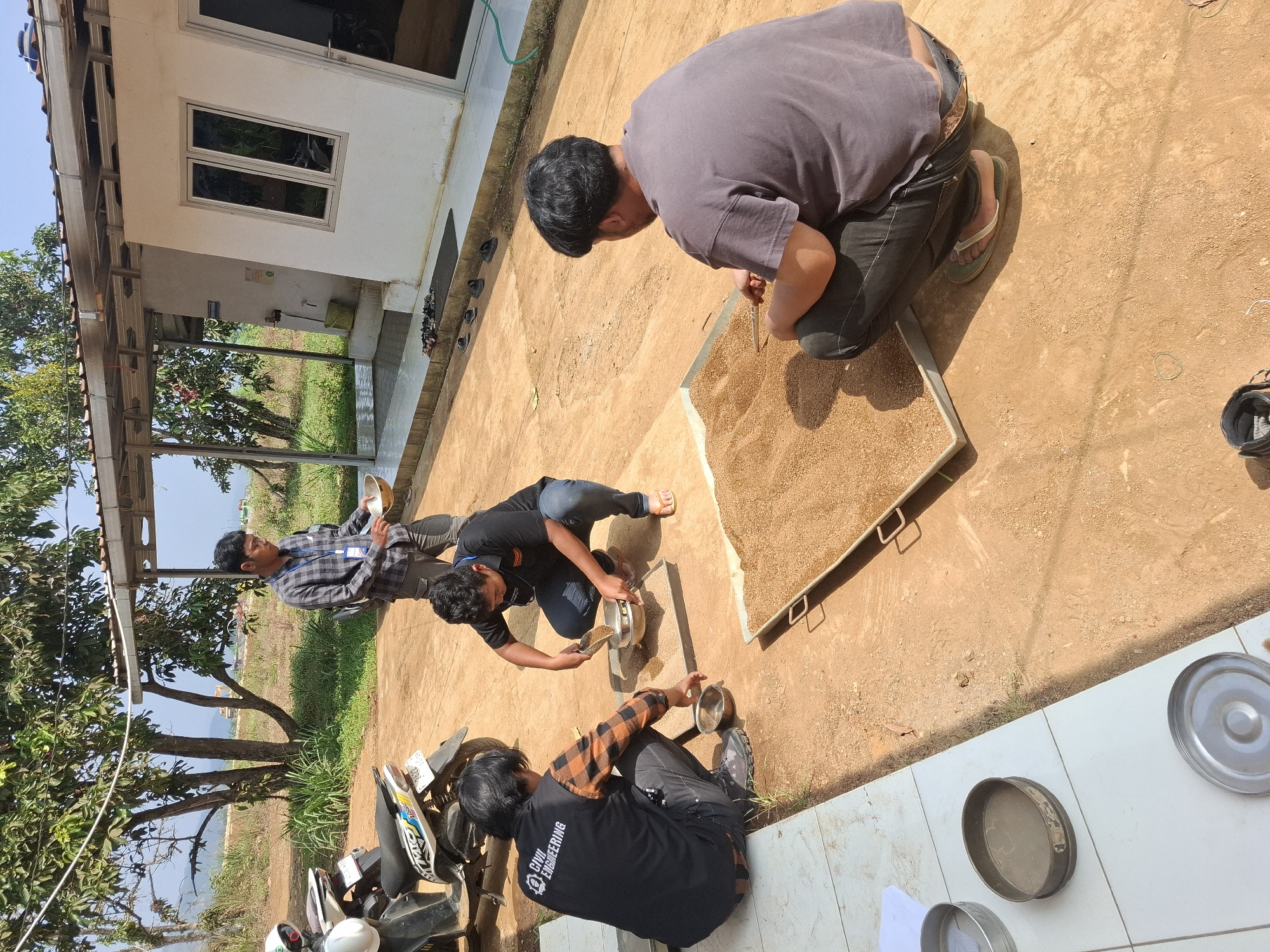 In Kutasirna, Indonesia, three individuals are engaged in a soil testing activity outdoors. One person, wearing a checkered shirt and dark pants, stands holding a metal bowl, looking down at the ground. Two other individuals are crouched down. One, in a black t-shirt with "CIVIL ENGINEERING" printed on it, is using a scoop to transfer material from a bowl into a sieve placed on a metal tray filled with fine particulate matter. The third person, wearing a dark grey t-shirt and black pants, is crouched beside a large metal tray also filled with the same granular material and is holding a wooden stick. The environment suggests a rural or semi-rural setting with a simple building with white walls and a corrugated metal roof in the background, surrounded by vegetation and trees. Two motorcycles are parked to the left of the frame. Several metal sieves and bowls are placed on a tiled surface in the foreground. The ground is composed of reddish-brown soil.