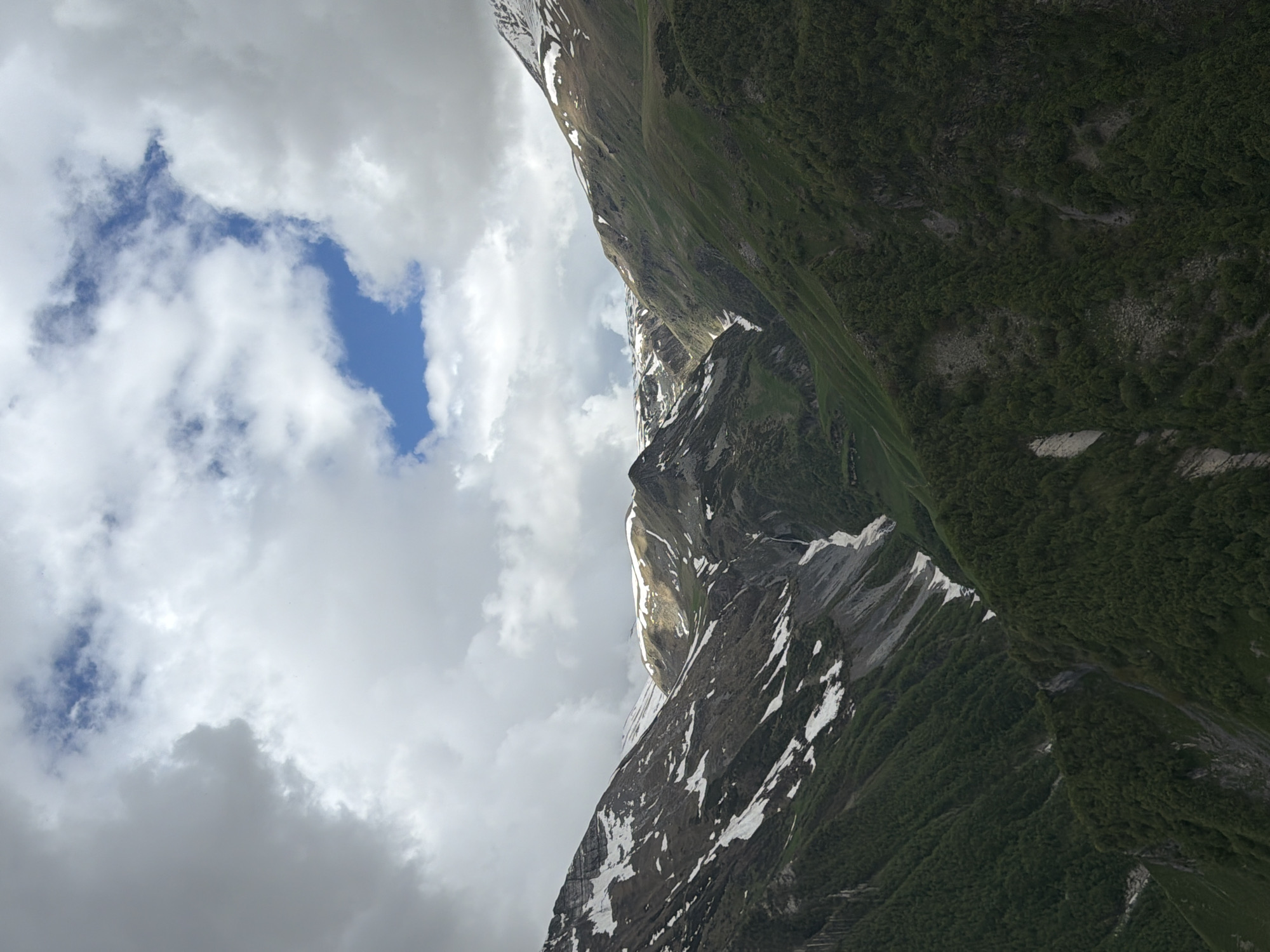 The image depicts a mountainous landscape, likely in Gudauri, Georgia, characterized by rugged terrain, verdant slopes, and patches of snow. The sky is overcast with dramatic clouds, allowing glimpses of blue sky. A narrow waterfall is visible cascading down a rocky incline. The lower slopes are densely covered with trees. The scene is natural and undisturbed, with no people or man-made structures evident. The lighting suggests it may be late afternoon or early morning, given the interplay of light and shadow on the mountainsides. The overall impression is one of a wild and majestic natural environment.
