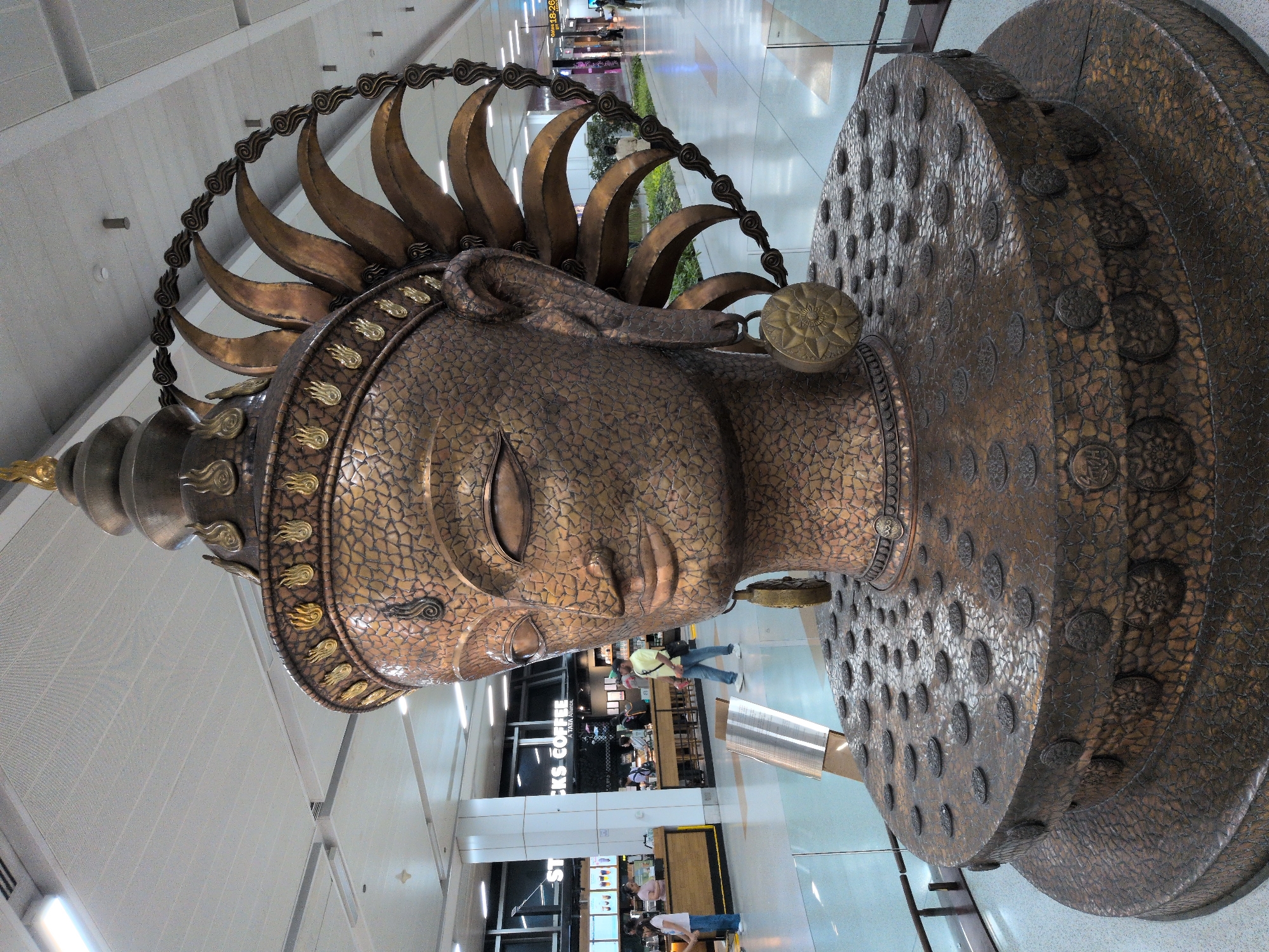 The image shows a large, ornate bronze sculpture of a head, likely of a deity or a significant religious figure, in an indoor public space, likely an airport terminal in New Delhi, India. The sculpture is detailed, with a textured surface on the face and head, and adorned with flames and a sun-like headdress. It sits on a multi-tiered base decorated with circular patterns. In the background, people are visible walking and in what appears to be a coffee shop with the "STARBUCKS COFFEE" logo clearly displayed. The environment is brightly lit with a modern ceiling and flooring, suggesting a contemporary public building. The sculpture is the primary subject and dominates the foreground of the image.