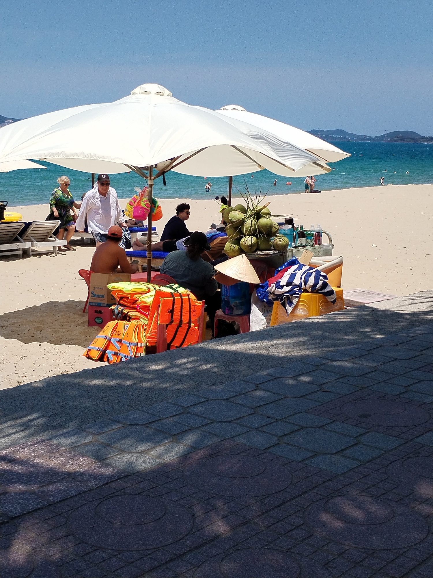 The scene is a beach in Nha Trang Ward, Vietnam, on a sunny day. The beach is light sand and there are several people in the water and on the sand. A large white umbrella shades a beach vendor stand. The stand has coconuts, bottled water, and orange life jackets. People are seated nearby. A shirtless man sits on a cardboard box with a logo that says "Sanna". A woman in a conical hat is present. A couple walks along the beach in the background. In the foreground, the patterned pavement is partially obscured by shadows.