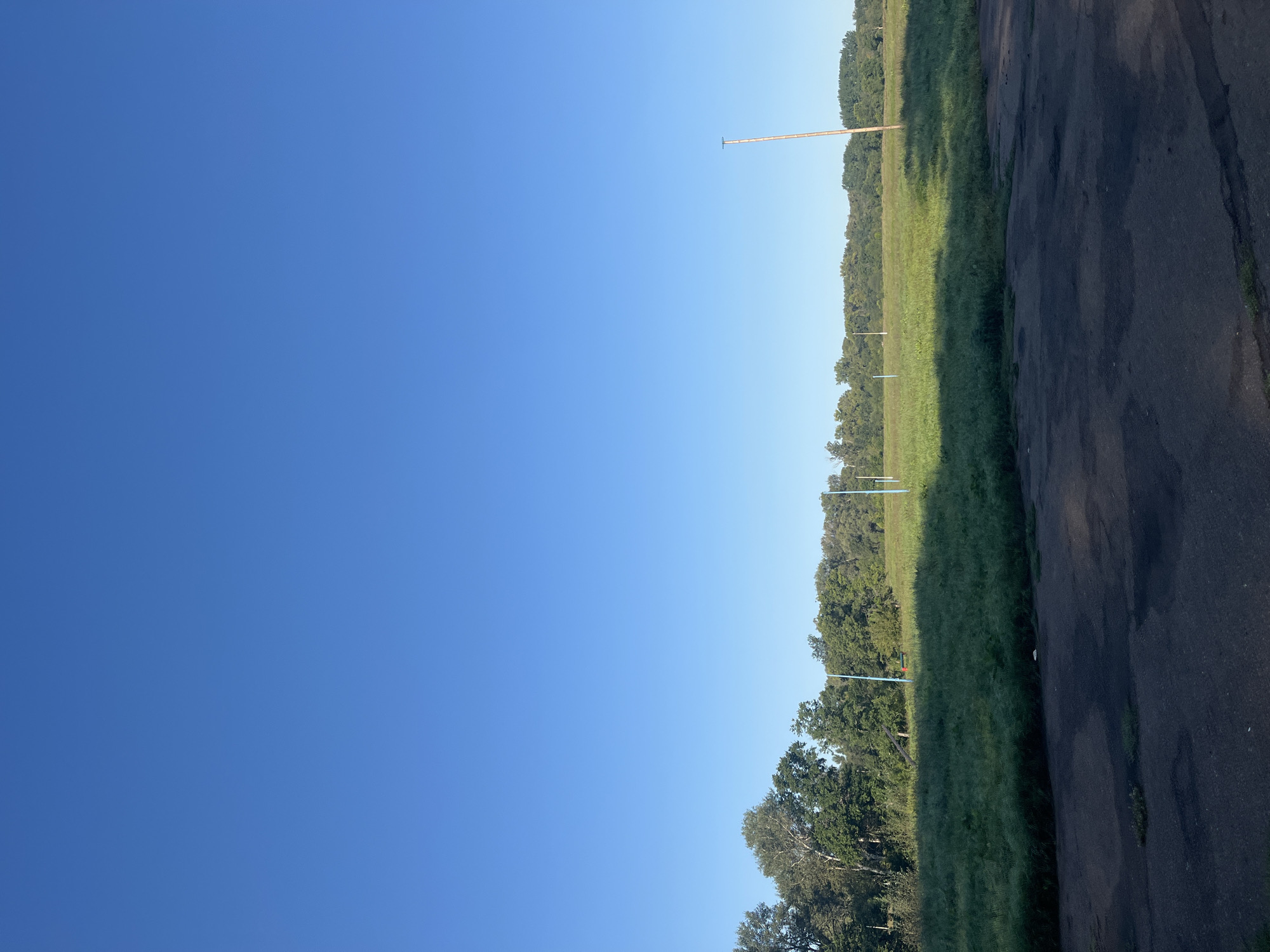 The image depicts a clear, bright blue sky stretching over a rural or park-like landscape. In the mid-ground, a dense line of green trees forms a horizon. In front of the trees, a grassy field is visible, with some shadows cast across it, suggesting the sun is at an angle. Several tall, thin poles are scattered across the field, with one prominent wooden pole on the right side of the image. Closer to the foreground, a dark, asphalt or paved surface with some cracks and patches of grass is visible. The scene appears to be an open, outdoor area, possibly a field or park on the outskirts of a populated area, given the presence of utility poles. There are no people or animals visible in the image. The overall impression is one of a tranquil, possibly early morning or late afternoon scene under a clear sky.