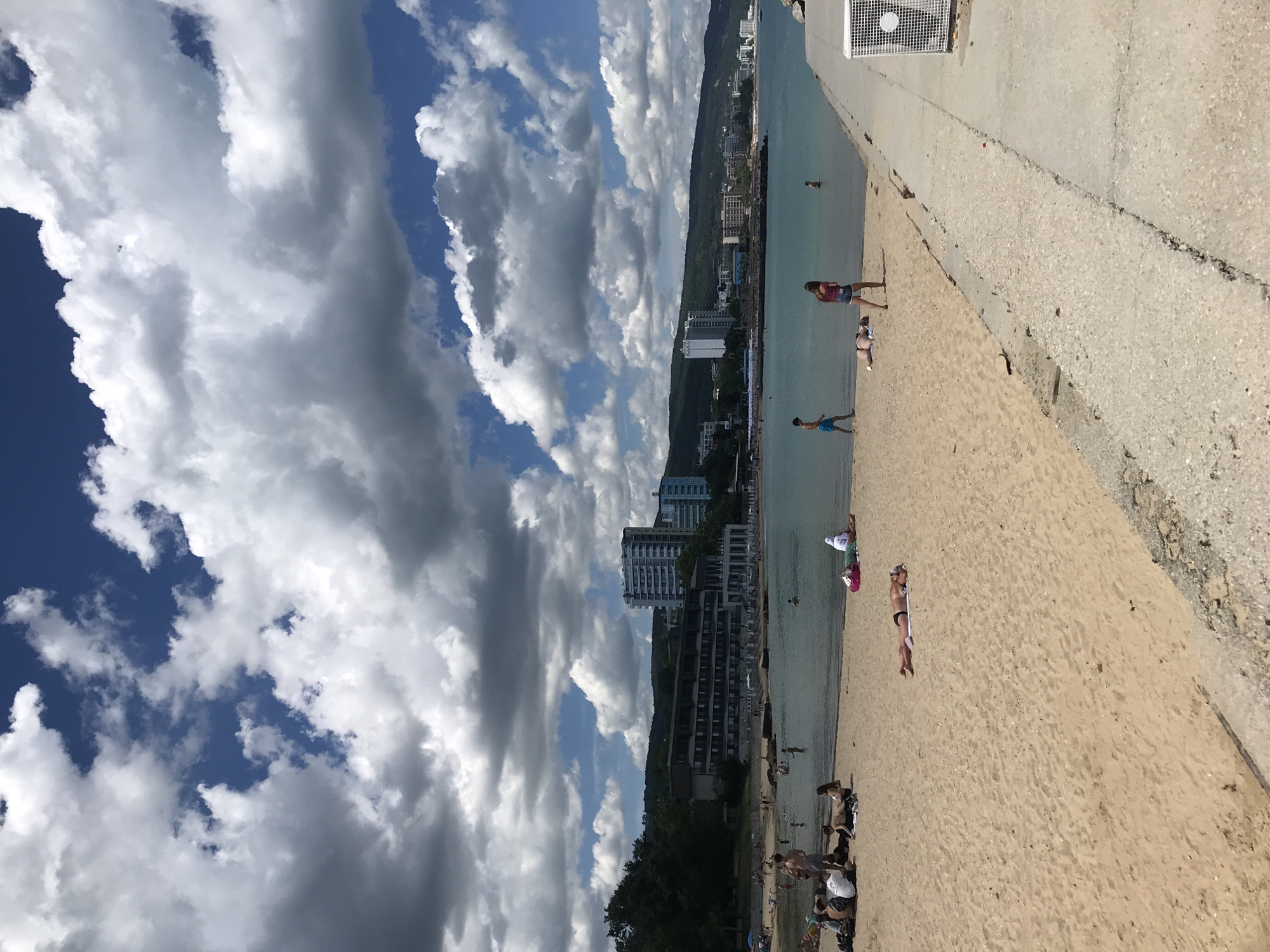 This image depicts a crowded beach scene on a sunny day with scattered clouds, likely on the coast of Varna, Bulgaria. The foreground is dominated by a sandy beach where several individuals are sunbathing and relaxing on towels. A man in dark swim trunks lies on his stomach, facing away from the camera. Further up the beach, a group of people are seated on towels, some appearing to be talking.
In the midground, people are in the shallow water, some wading and others swimming. The water is a clear, calm turquoise. Beyond the water, a strip of coastline is visible, lined with trees and several multi-story buildings, including hotels and apartments, characteristic of a resort town. The most prominent structures are modern, light-colored high-rise buildings. The Black Sea stretches out to the horizon, meeting a bright blue sky filled with dramatic white and gray cumulus clouds. A concrete walkway or pier runs along the right side of the image, with a white ventilation unit visible at the far end. The overall impression is a lively summer day at a popular beach destination.