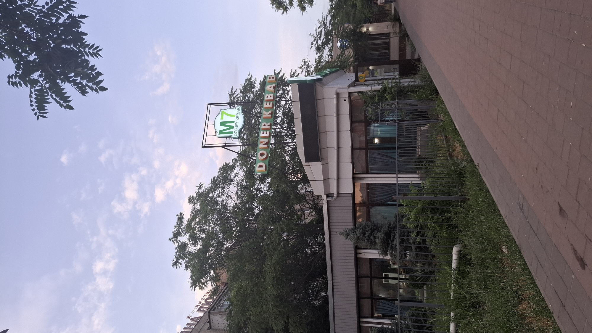 The image shows a street scene in Shymkent, Kazakhstan. A prominent sign for "M7 DONER KEBAB" is visible, illuminated with green and white lettering. The sign is mounted on a pole in front of a modern building with large windows, some of which are covered with curtains. Trees and greenery surround the building, with a paved walkway in the foreground. The time of day appears to be late afternoon or early evening, as the sky is a soft blue with scattered clouds, and the lighting suggests it is not yet dark. There are no people visible in the image. The overall impression is that of a commercial establishment on a quiet street.