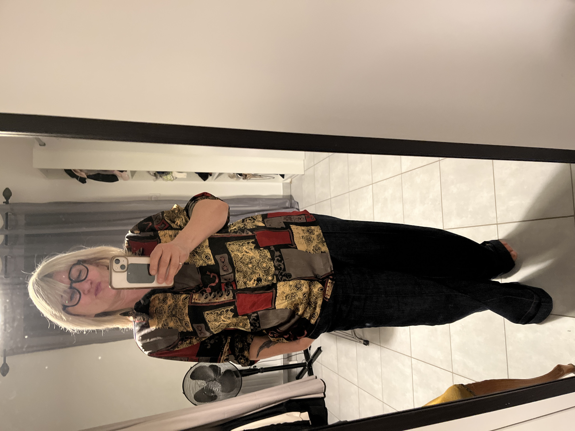 A mirror selfie captures an individual indoors in Chambœuf, France. The subject, a blonde-haired person wearing dark-rimmed eyeglasses, stands facing the mirror with puckered lips. They are dressed in a patterned long-sleeved shirt featuring red, gold, black, and grey sections, paired with dark blue wide-leg jeans. Their left hand is placed in their pocket, and their bare feet are visible. Their right hand holds a light-colored smartphone, indicating the image capture method. A tattoo is visible on the subject's left forearm, and a ring is present on the ring finger of their right hand.
The reflected environment features light-colored tiled flooring. To the left of the subject in the reflection, a black pedestal fan and a pale grey curtain are visible. An open shelving unit or wardrobe containing various clothing items is visible on the right side and behind the subject, set against a light-colored wall. A dark curtain rod with finials is present at the top of the reflected background. The mirror itself has a dark, thin frame along its right edge.