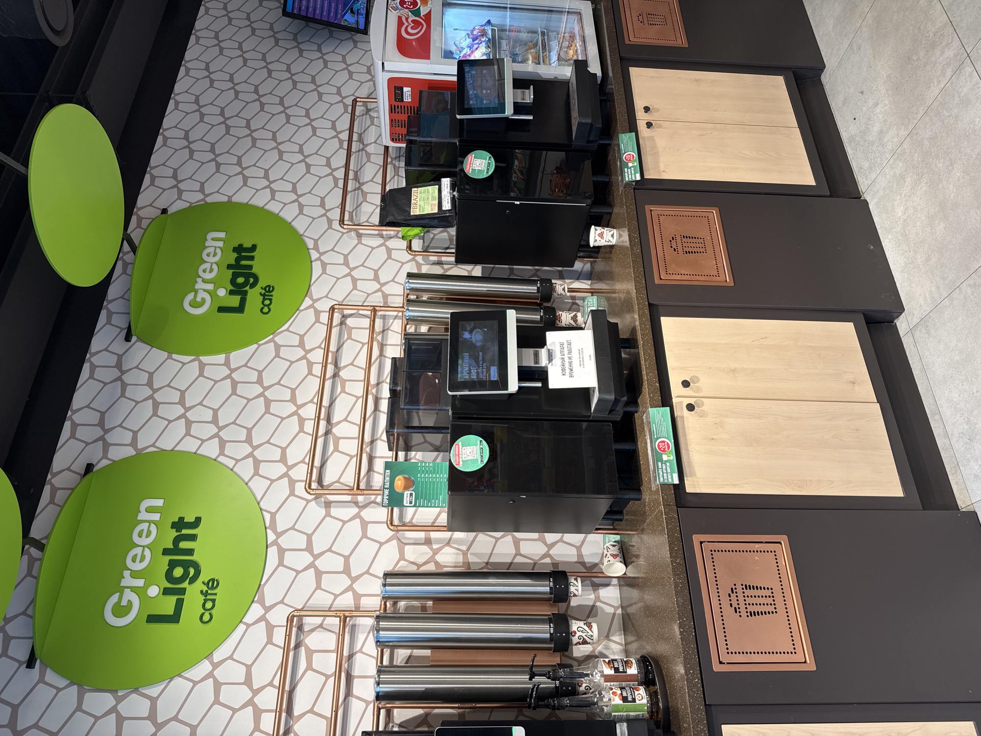 The image shows a "Green Light Café" counter in Medovoye, Russia. Two lime-green circular signs with the café's name are visible on the left, adjacent to two dark-green chairs. Behind the signs are two sets of coffee machines, each with a screen, copper-colored metal frames, and coffee-making components. Bags of "BRAZIL" coffee are visible on one of the counters. One of the counters displays a printed sign that reads "COFFEE MACHINE IS TEMPORARILY NOT WORKING". To the right of the machines are dark-brown countertops with light-wood-colored cabinet doors, and copper-colored metal squares with hole patterns are installed. A vending machine is also partially visible in the background. The floor has a honeycomb pattern in light-gray and white tones. The lighting is bright and fluorescent.
