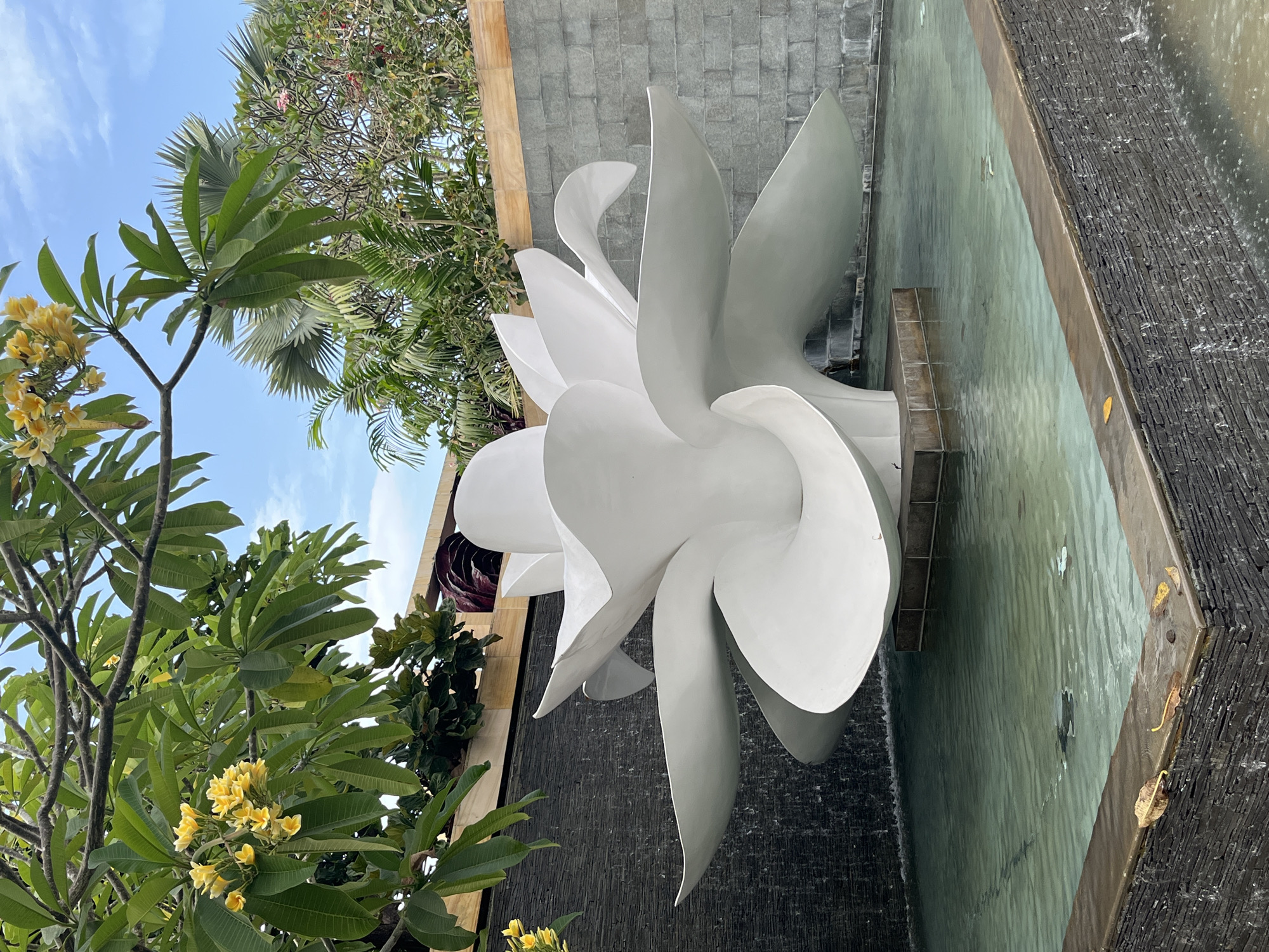 A white, sculptural lotus flower stands in a shallow, rectangular pool of turquoise water. The sculpture is positioned in the center of the pool, with its layered petals fanning outwards. The pool is surrounded by dark gray, stacked stone walls, with a darker, textured stone wall behind it, from which water appears to be cascading down. Lush green foliage, including frangipani trees with yellow blossoms, and palm trees, frame the scene against a bright blue sky. The overall impression is one of a serene, resort-style outdoor setting, likely in Kuta Selatan, Indonesia, a popular tourist destination known for its beaches and hotels.
