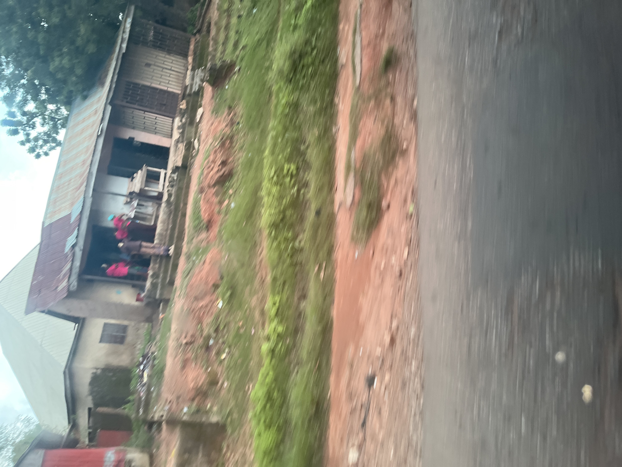 The image depicts a street scene in Akwanga, Nigeria, showing a weathered building with a corrugated metal roof and a partially visible red structure to the left. Several people, including a person in a bright pink garment and another in a brown outfit, are visible near the building's entrance, suggesting activity. The ground in front of the building appears to be red earth, with patches of green grass and some debris. Further ahead, the terrain slopes upwards, with more red earth and sparse vegetation. The foreground is dominated by a blurred grey surface, likely a road, with some white and yellow smudges that could be reflections or debris. The overall impression is that of a rural or semi-urban environment with simple architecture and a somewhat unkempt appearance.