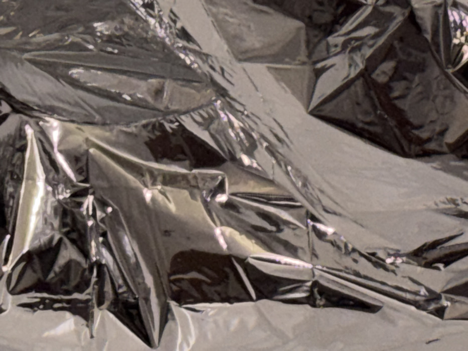 The scene depicts an abstract, close-up view of a highly crumpled, reflective metallic sheet, likely aluminum foil. The material exhibits numerous sharp creases, folds, and undulations across its entire visible surface, creating a complex, fragmented pattern of bright specular highlights and deep shadows. The predominant color palette consists of varying shades of silver, grey, and black, with subtle, faint chromatic shifts visible as light reflects off the textured surfaces. No discernible people, subjects, objects (other than the foil), actions, or interactions are present within the frame. The environmental context is limited to this material. This scene is located in Tunbridge Wells, United Kingdom.