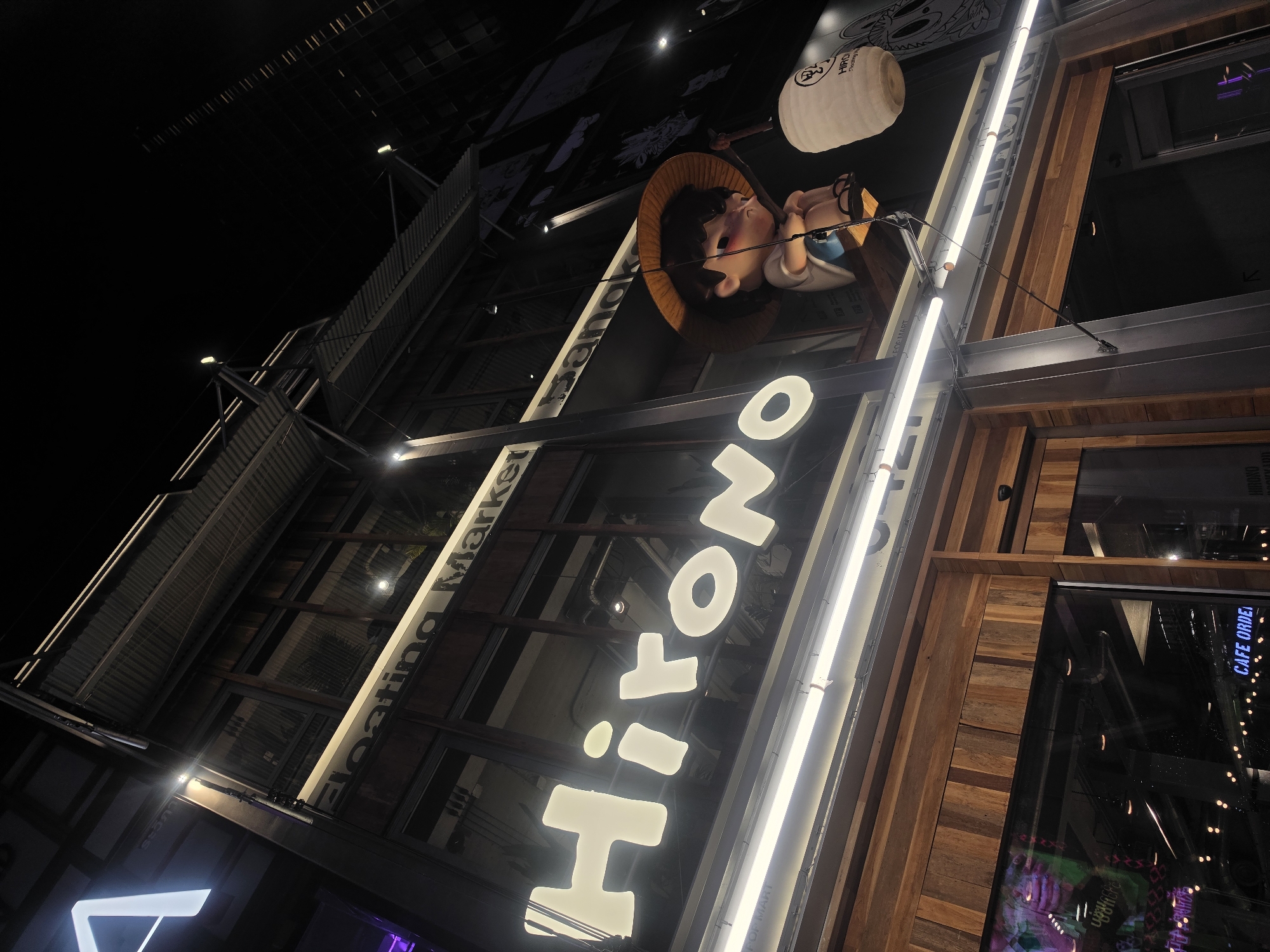 Captured from a low angle at night, the image showcases the exterior of the "Hirono Floating Market" in Bangkok, Thailand. A large, illuminated vertical sign spells out "Hirono" in stylized, white lettering. Above the sign, a cartoon figure of a person wearing a straw hat and glasses holds a lantern. The building is constructed with a mix of metal, glass, and wooden elements. A brightly lit fluorescent lamp runs vertically along the right edge of the building. The interior, partially visible through the windows, has lights that appear to be cafe or restaurant decorations.